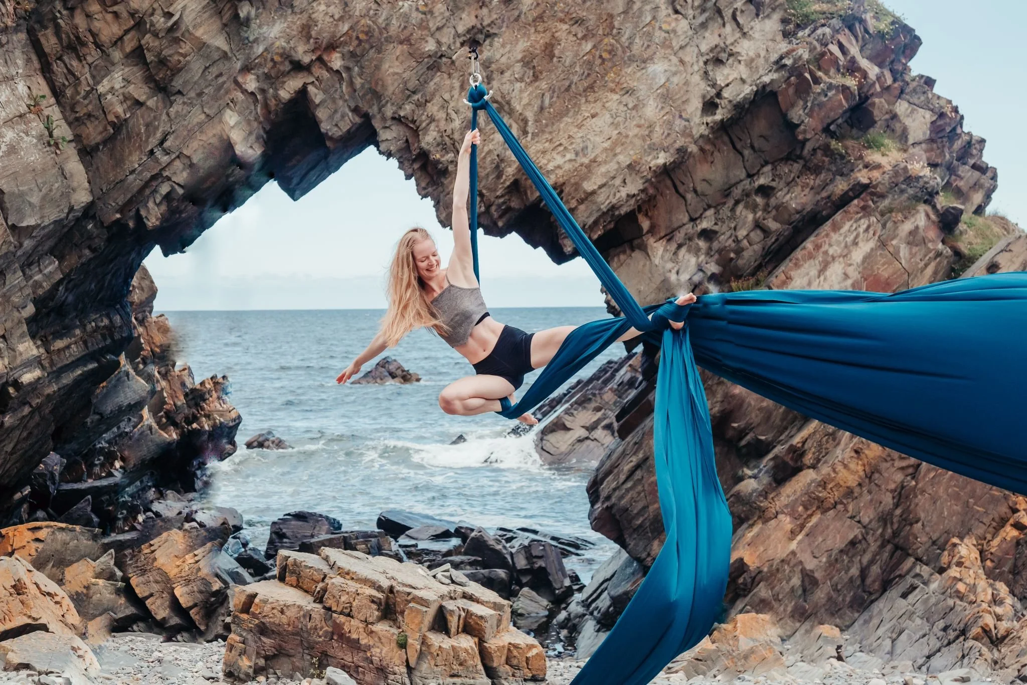 A woman practicing aerial silks on blue fabric suspended between rocky cliffs by the seaside