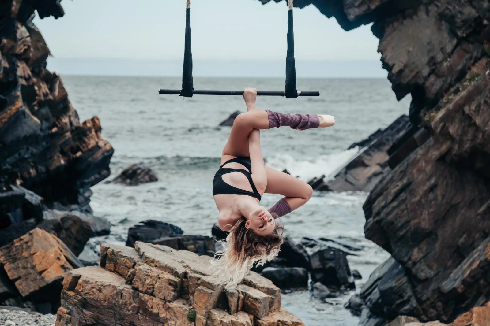A woman in a black performance outfit  and striped thigh-high socks is hanging upside down from a horizontal bar between rocks by the beach, with wavy blonde hair and an ocean in the background.