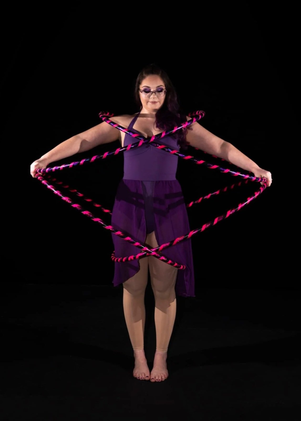 Woman in purple dress holding multiple pink and black hula hoops in front of a black background.