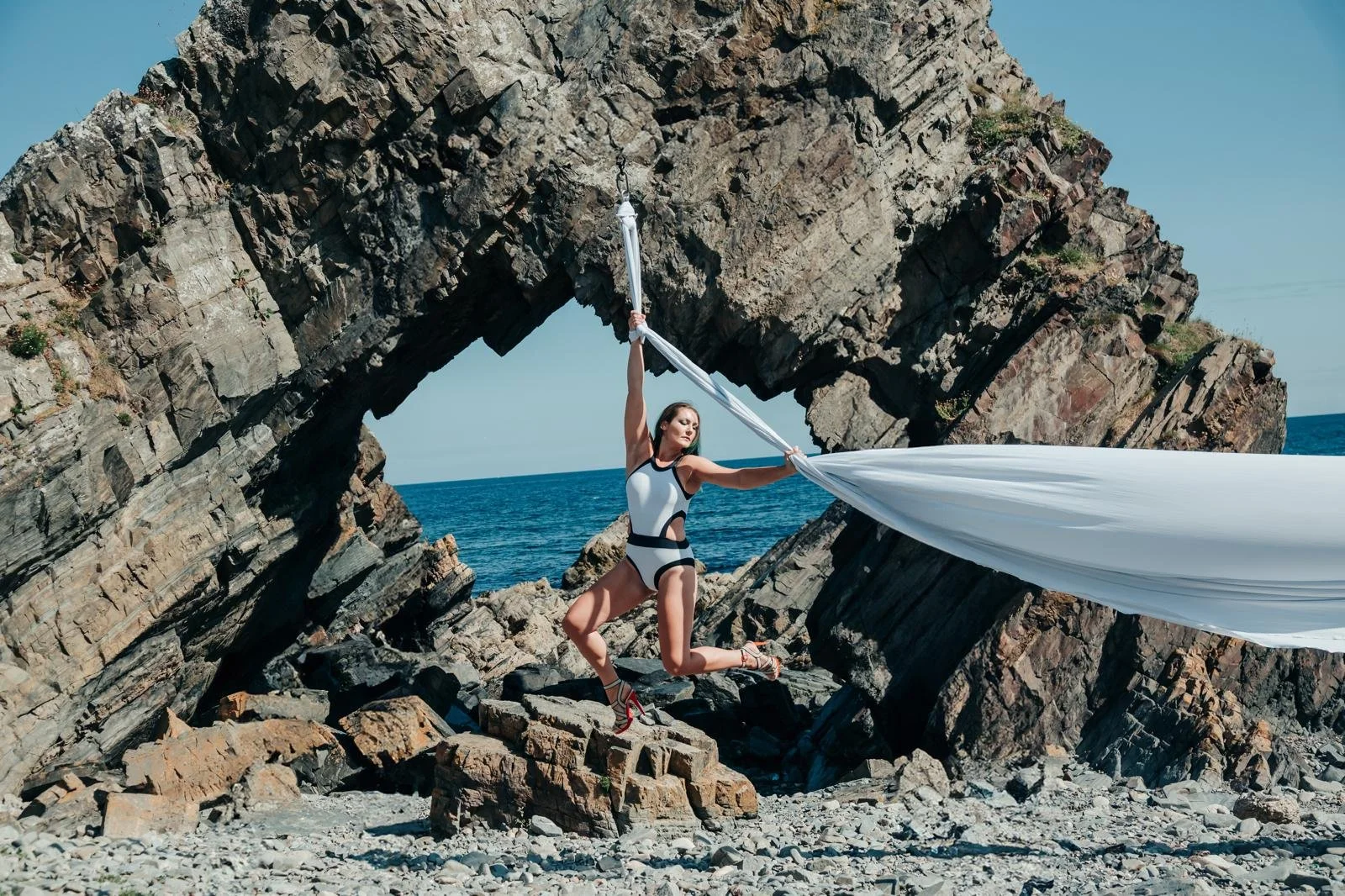 A woman in a black and white performance outfit standing on rocky shore, holding a long white fabric, with a large rocky arch formation and ocean in the background.