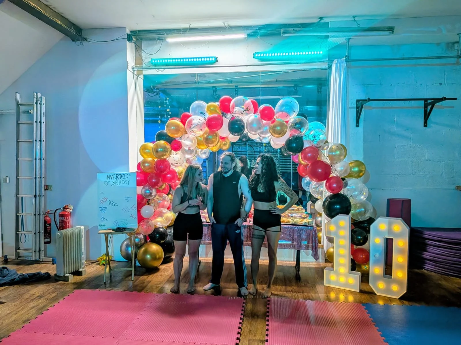 Two women and one man stand beneath a balloon arch at a celebration, with large illuminated numbers '10' on the right, on a pink and blue mat. The background features a table with decorations and a whiteboard mentioning a birthday.