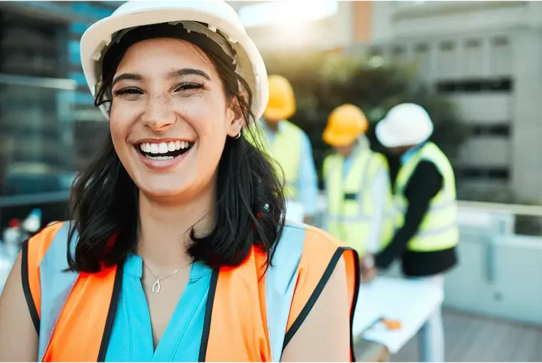 A smiling woman in a construction helmet and safety vest at a construction site with other workers in the background.