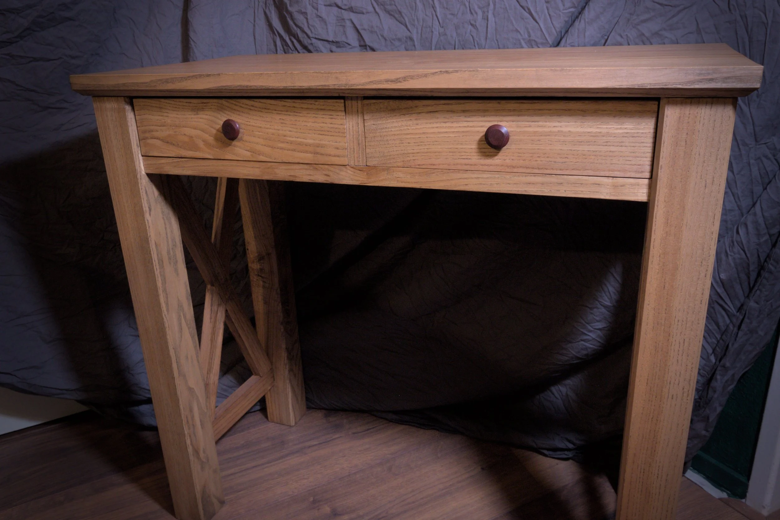 Een houten tafel met twee laden en een kruisvormige ondersteuning aan de zijkant, geplaatst op een houten vloer met een zwarte stoffen achtergrond.