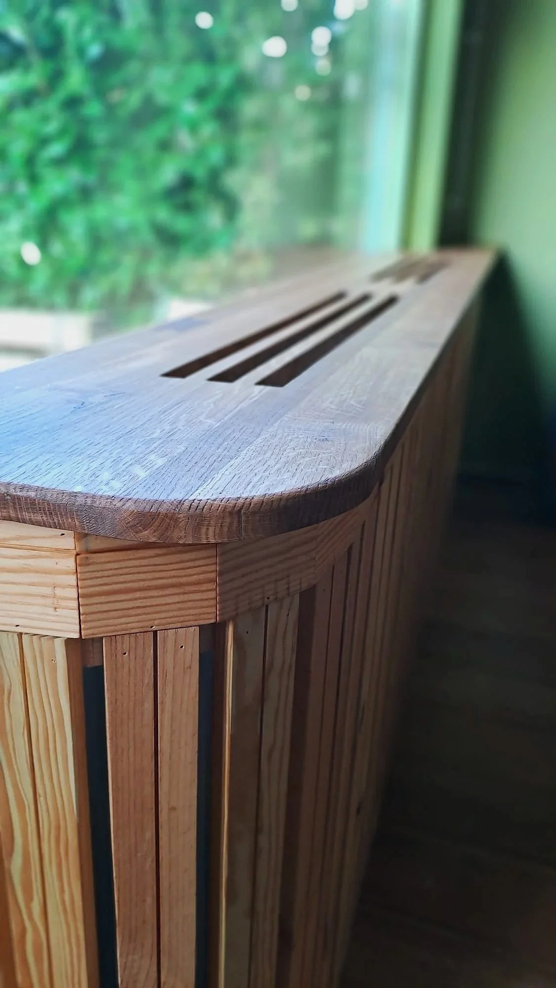 Houten radiatorombouw met lange, dunne houten latten en een gebogen rand naast een raam met groen uitzicht.