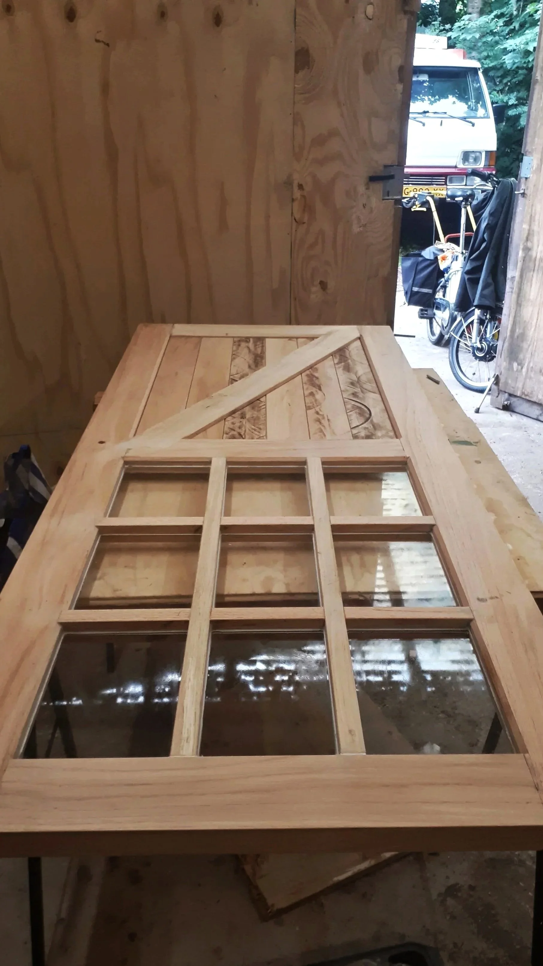 Een houten paneeldeur met ramen en vakjes, ligt op een werktafel in een houtwerkplaats.
