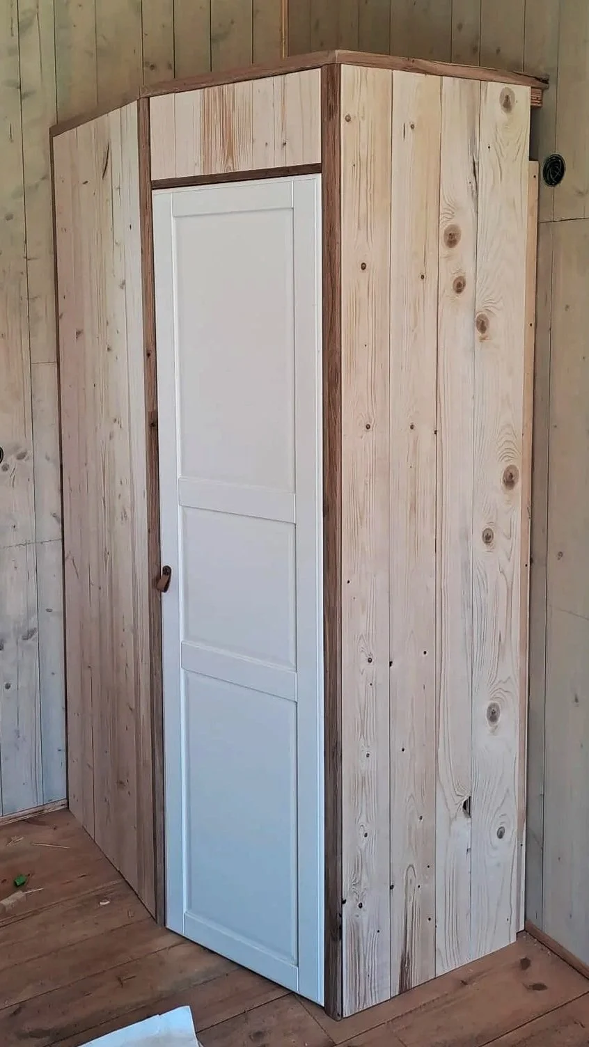 Een houten kast in een kamer met houten wandpanelen en houten vloer, de kast heeft een witte deur.