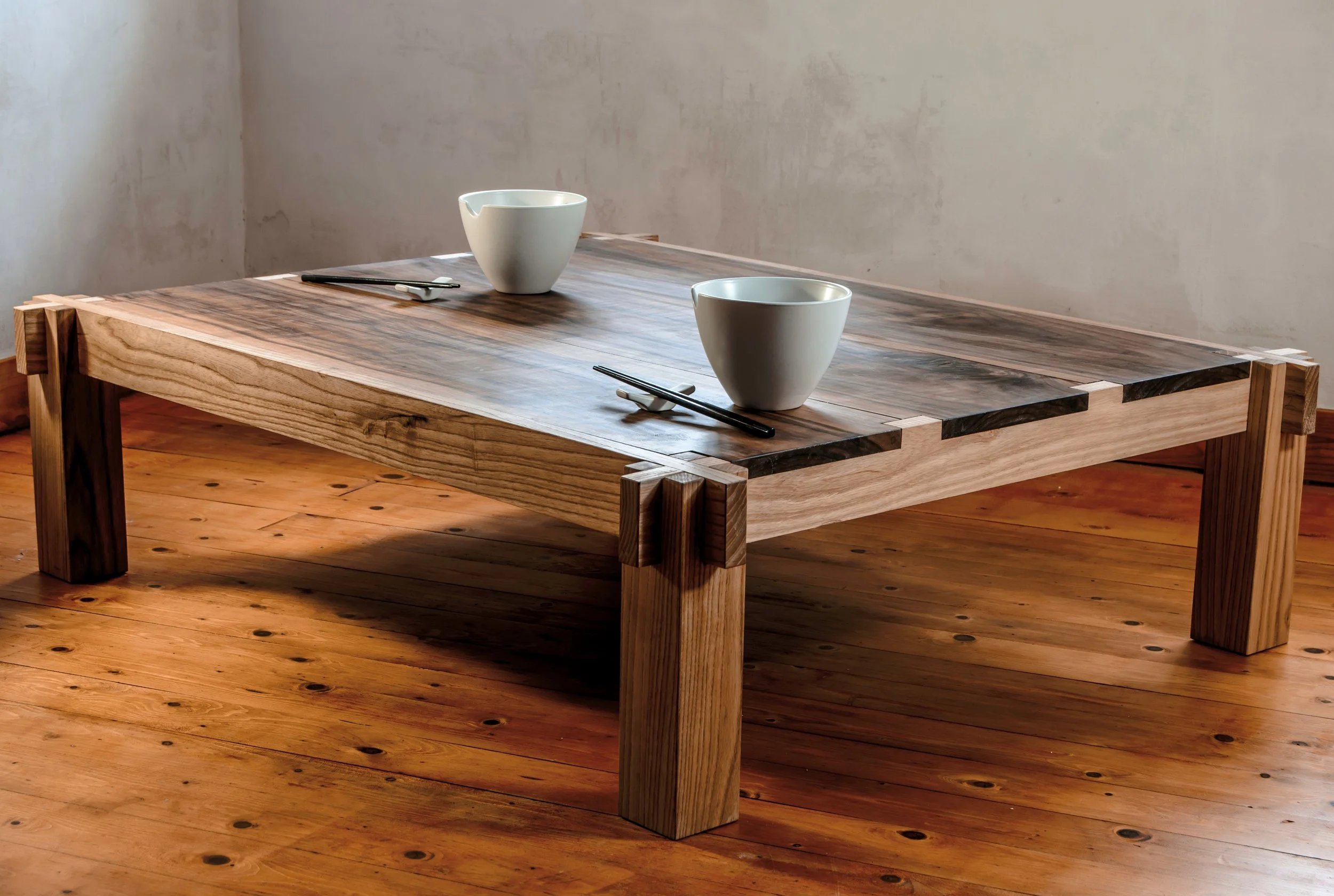 Een lage houten tafel met een rustieke uitstraling, geplaatst op een houten vloer, met twee witte kommen en bestek erop.