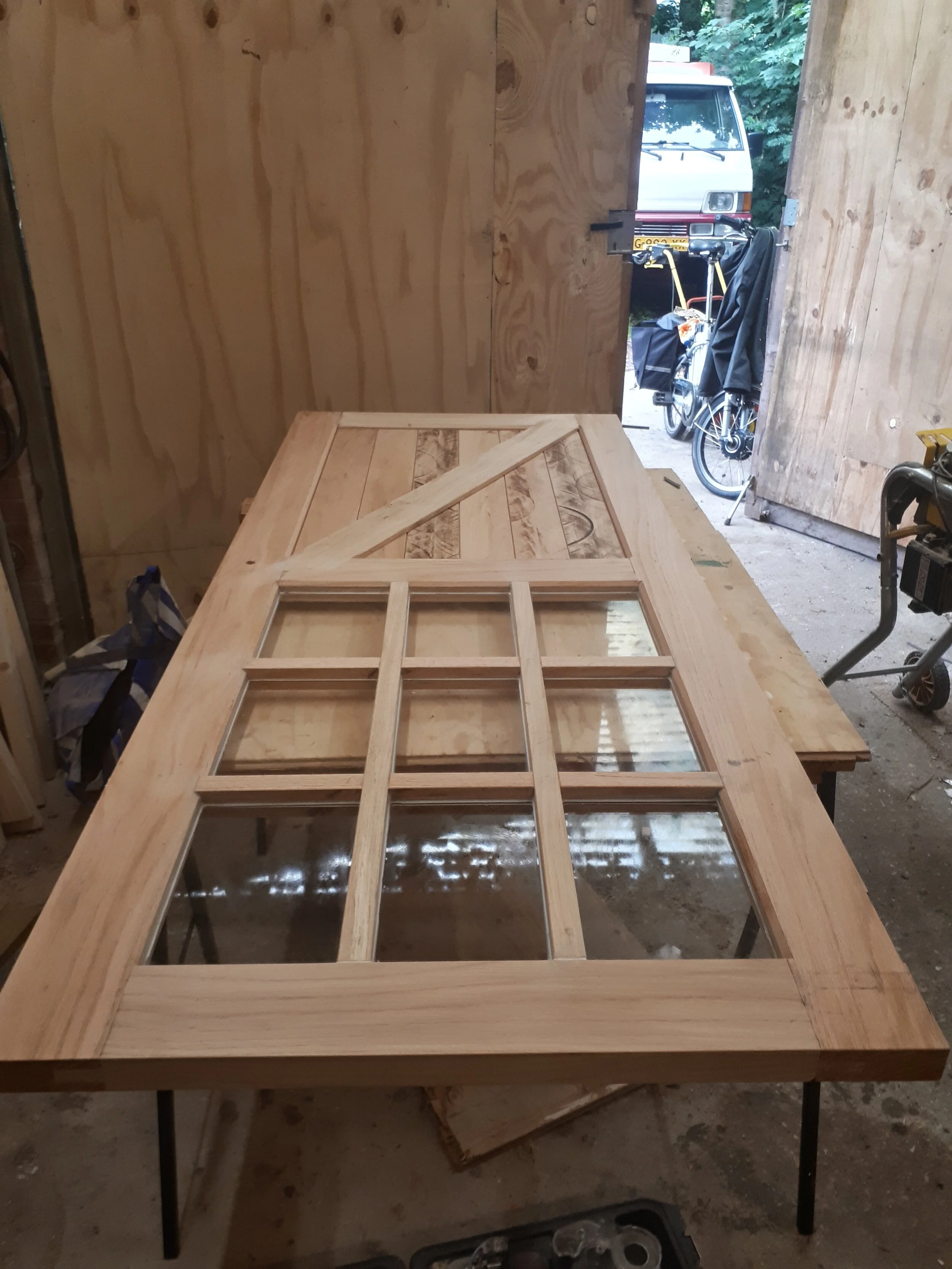 Een houten deur met ramen onder constructie, gelegen op een werkbank in een werkplaats.