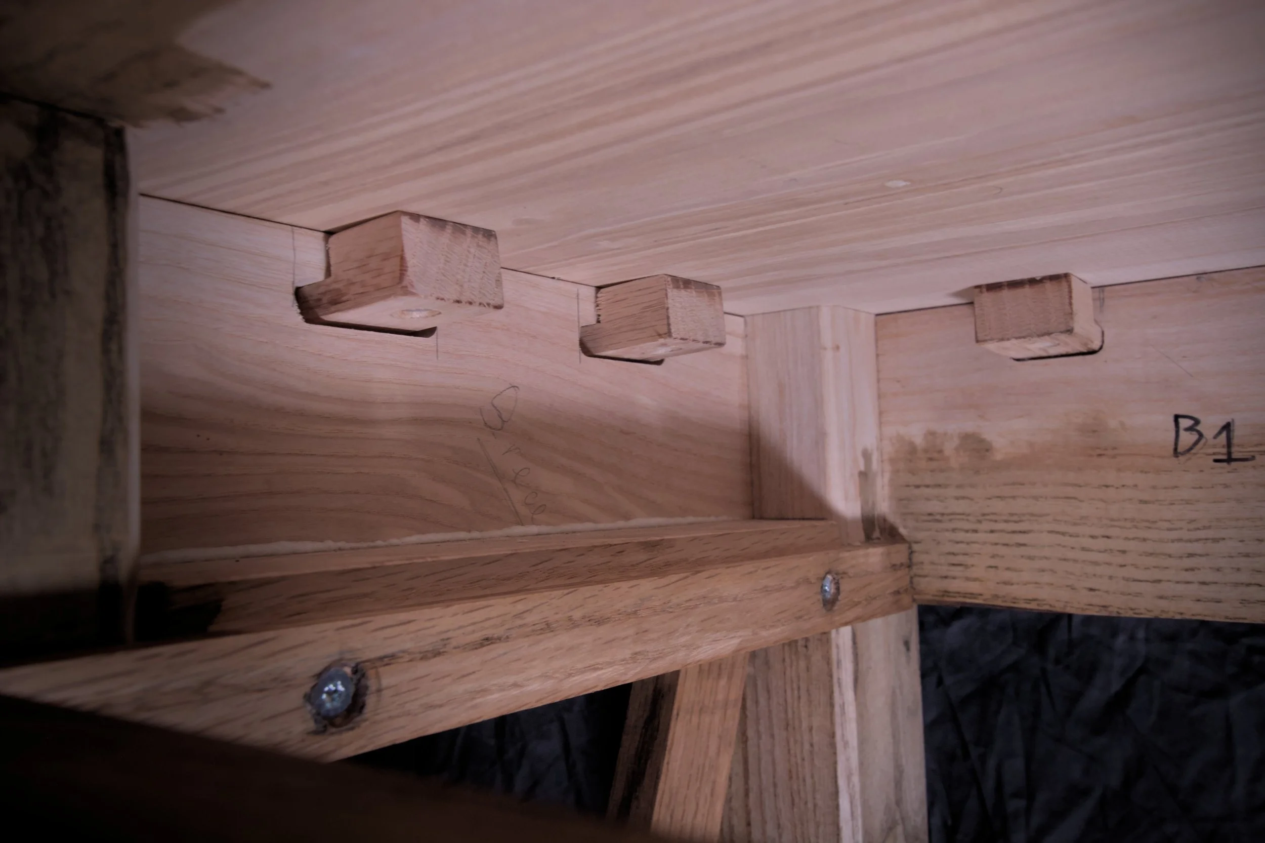 Binnenkant van een houten meubelstuk met drie houten wiggen en schroeven, duidelijke houtbewerking, en enkele handgeschreven markeringen.