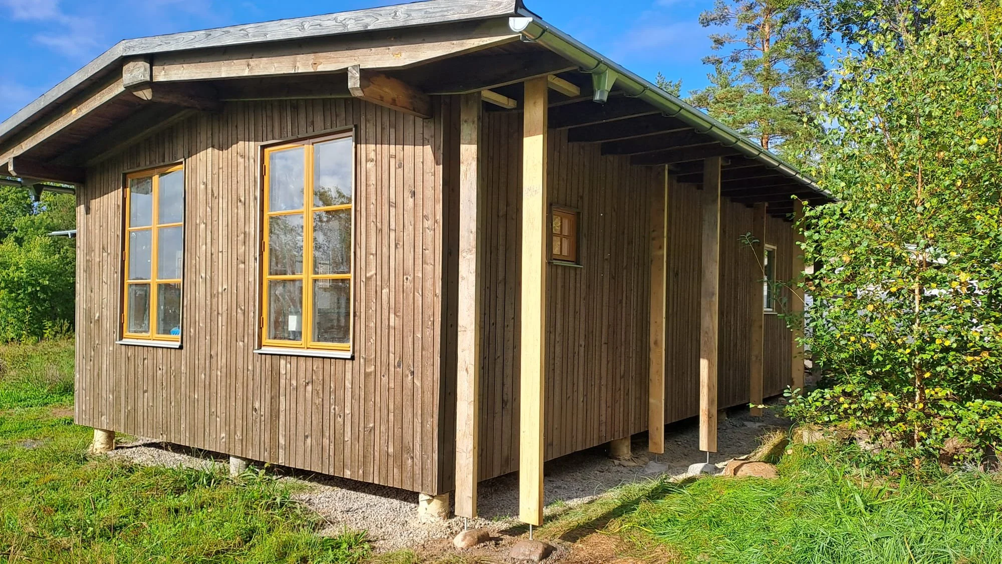 Houten huis met houtopslag in aanbouw drie grote ramen en een zijdeur, omgeven door groene planten en bomen.