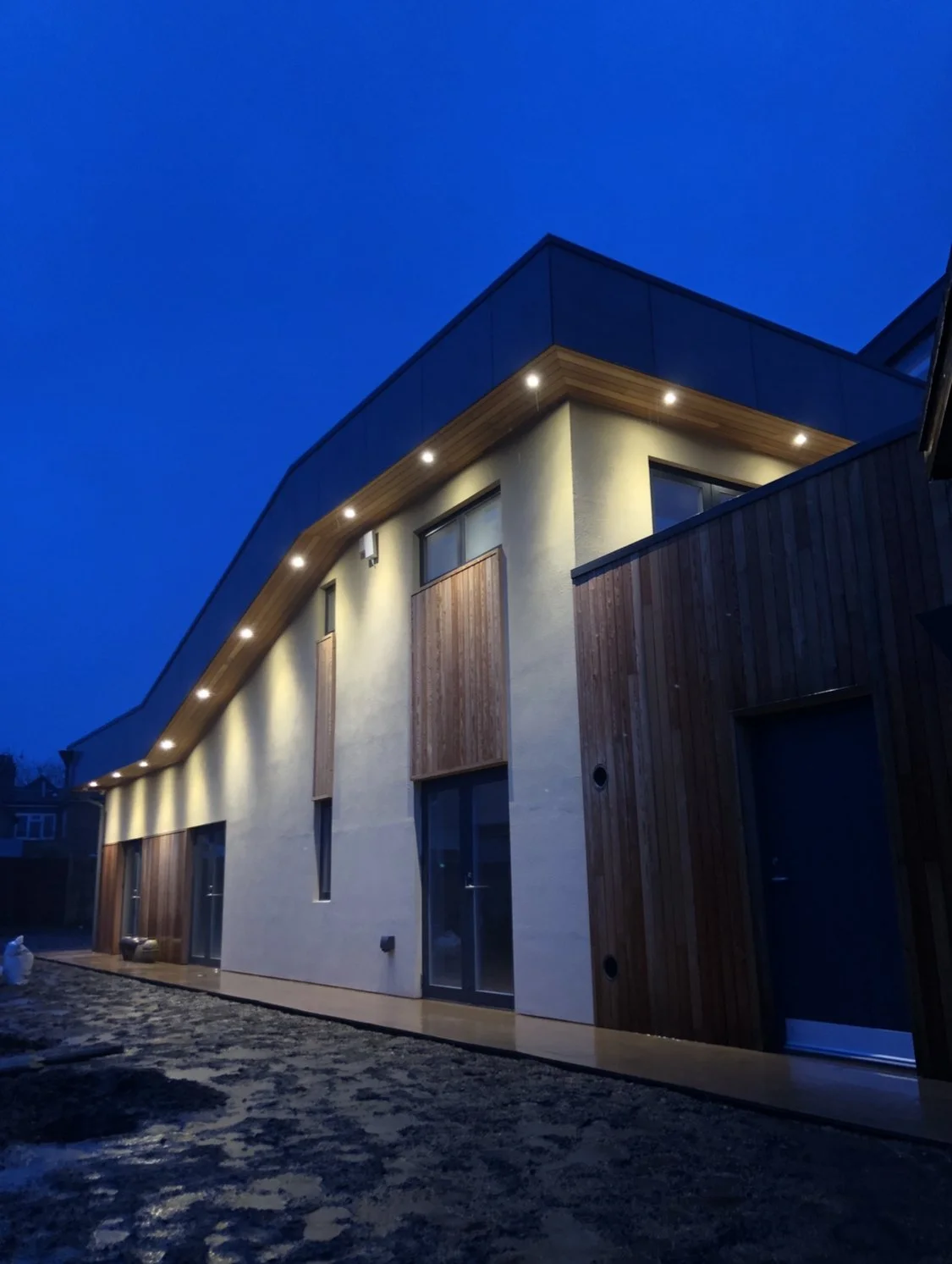 Modern multi-story house with wooden and white exterior walls, illuminated by exterior lights at dusk, with a dark sky in the background.