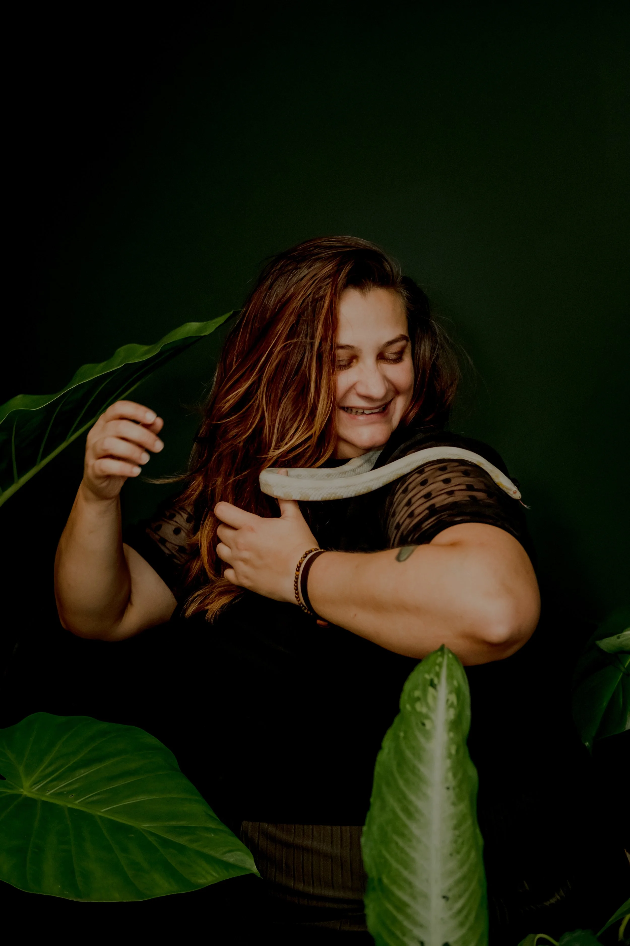 A smiling woman with long brown hair and tattoos, holding a white snake around her shoulders, surrounded by large green leaves, against a dark green background.