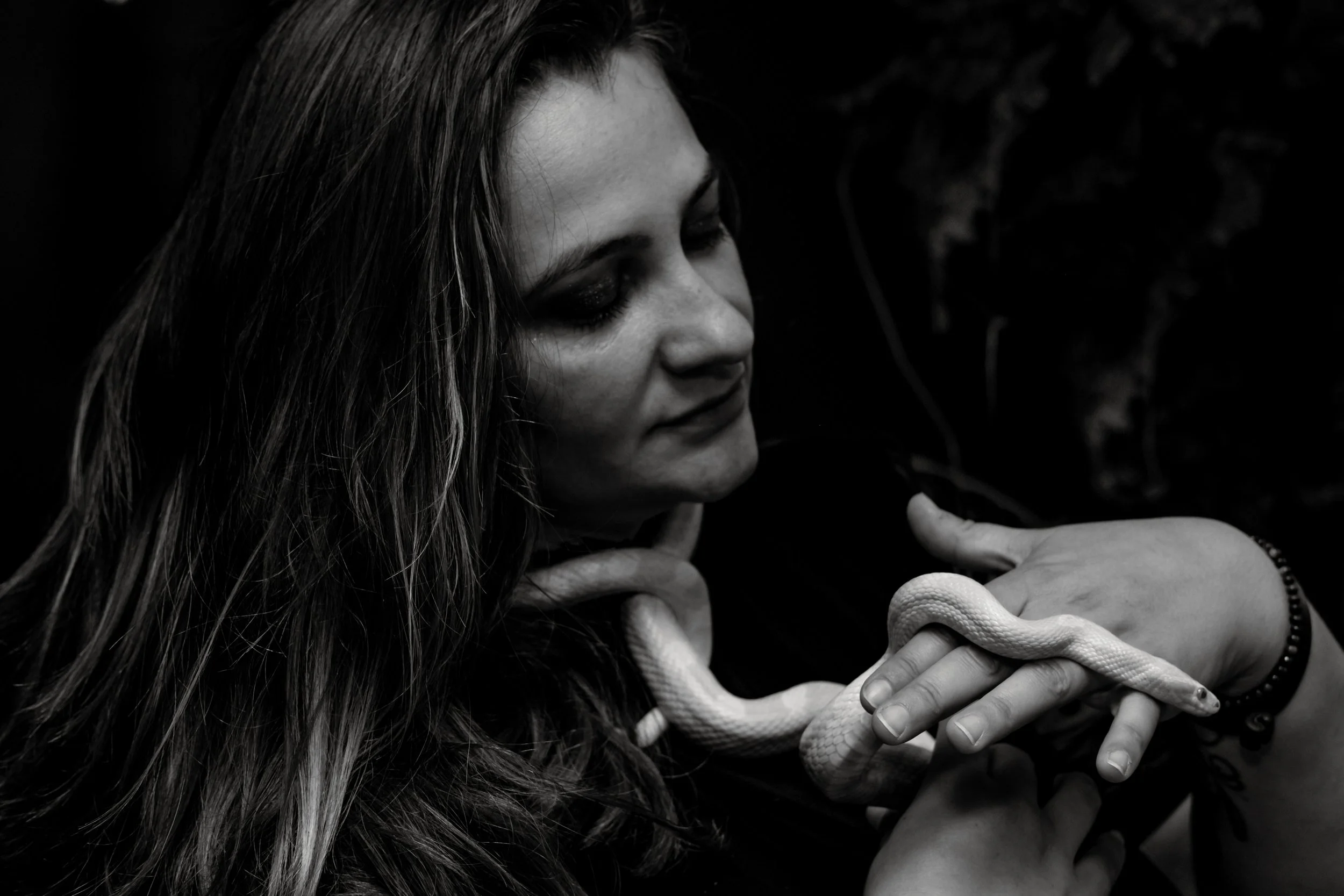 A woman with long hair, dark eye makeup, and a black shirt holds a white snake, gently holding it in her hand with a calm expression, against a dark background.
