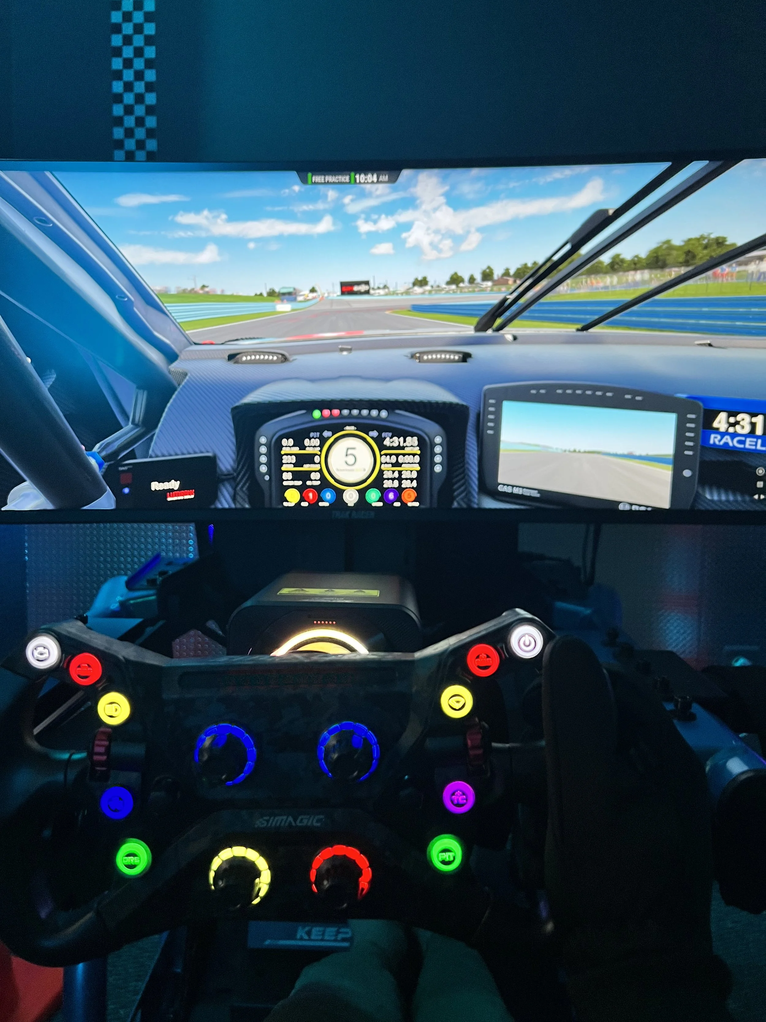 Inside a race car cockpit at a racetrack, with steering wheel, digital displays, and a view of the racetrack ahead under blue skies with scattered clouds.