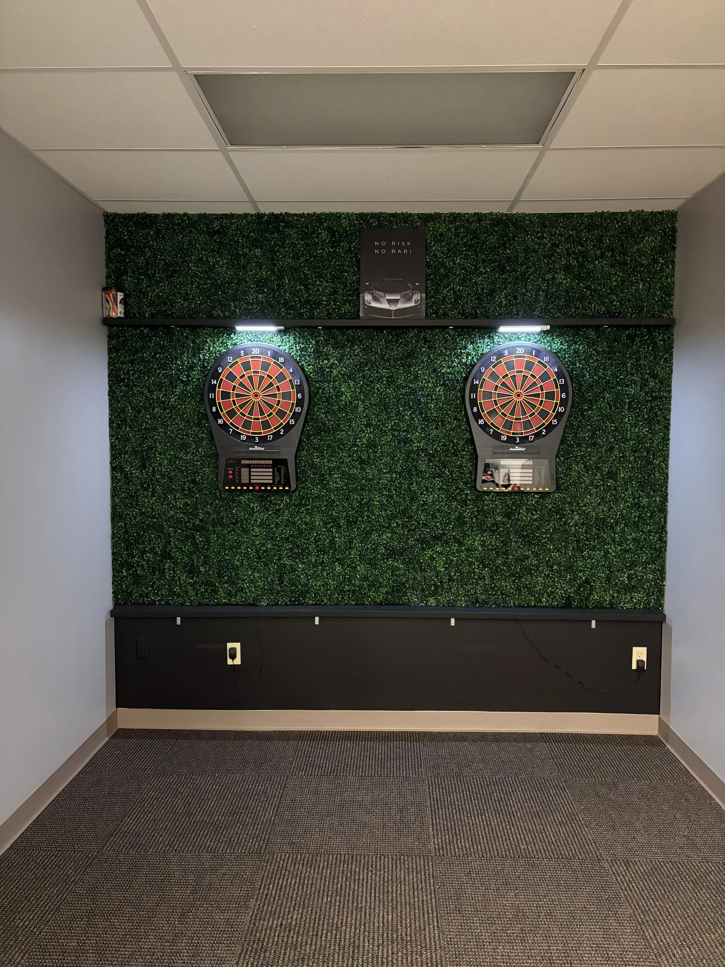 Two electronic dartboards mounted on a green leafy textured wall, with a small black shelf above them and a poster of a sports car in the middle. The floor is carpeted with a dark patterned carpet.