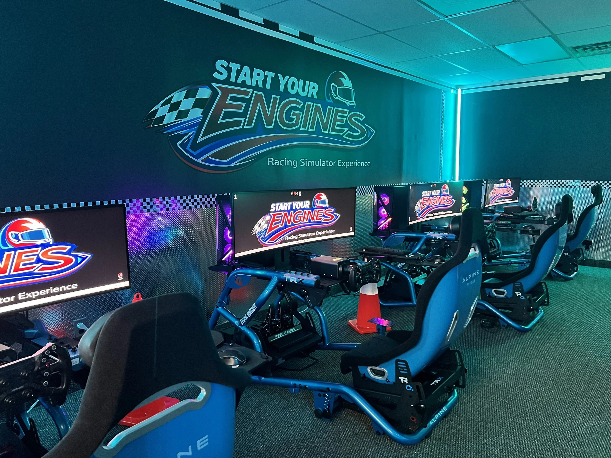 Indoor racing simulator experience with multiple gaming stations, each equipped with racing seats, steering wheels, and monitors, under a neon-lit sign that reads "Start Your Engines".