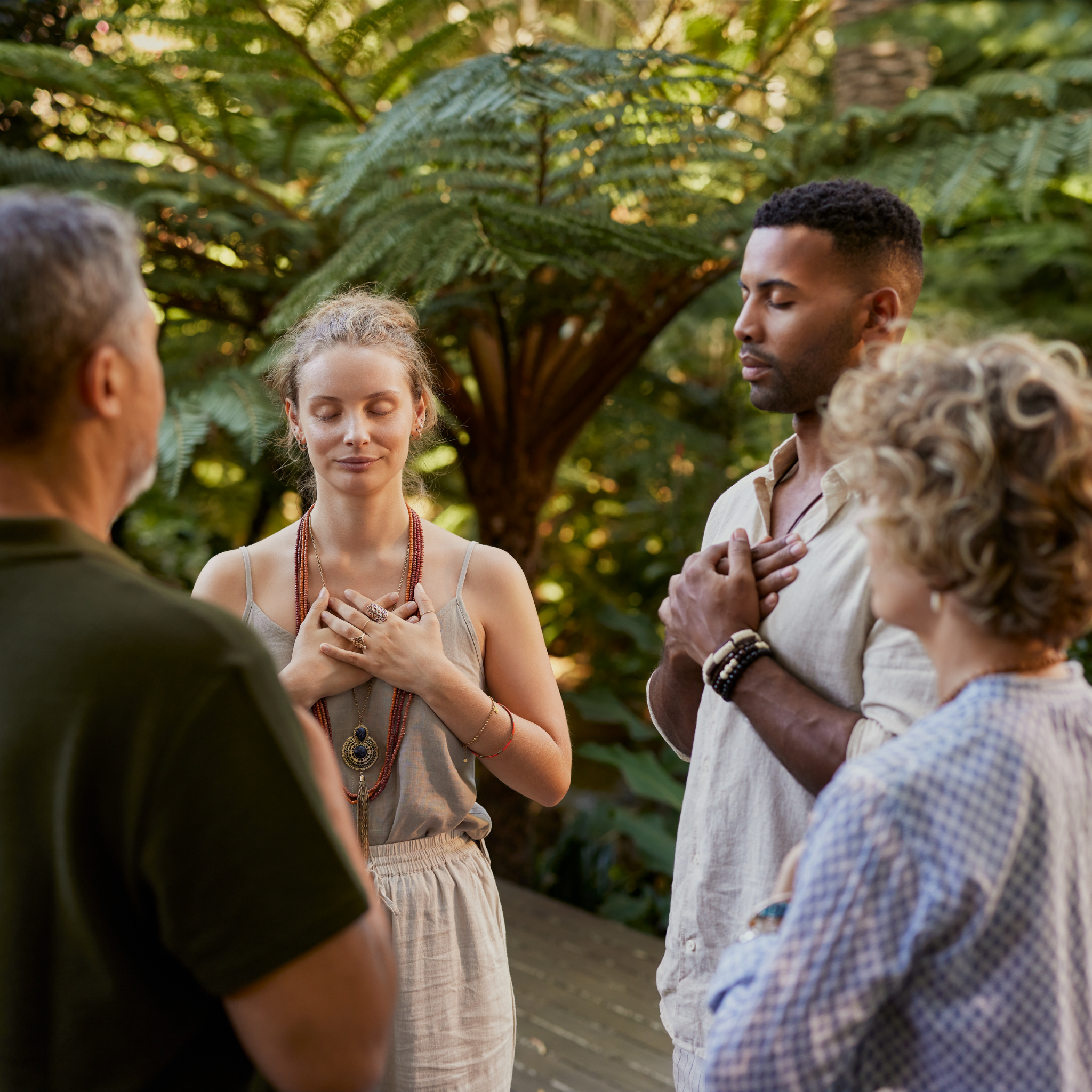 People standing in a circle outdoors, with one woman in the center with her eyes closed and hands over her chest, engaging in a meditation or prayer, surrounded by lush green foliage.
