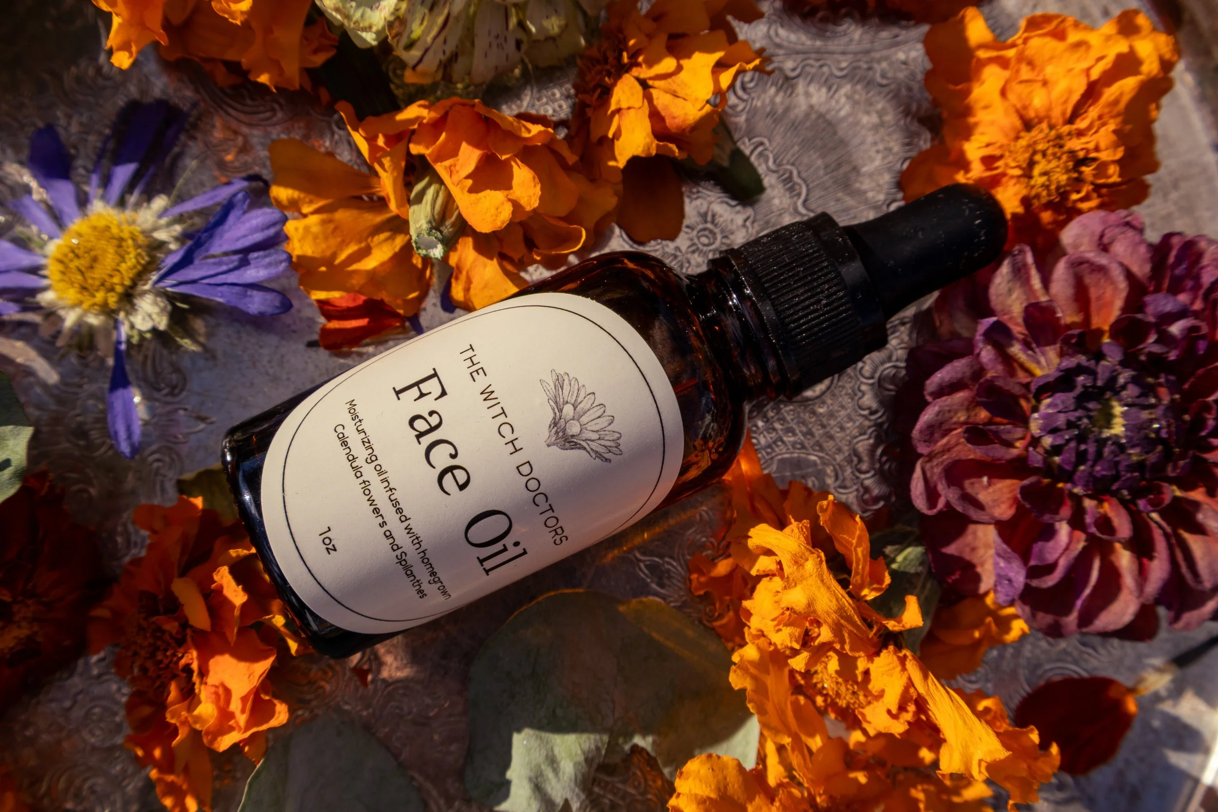 A bottle of face oil surrounded by various colorful flowers on a textured surface.