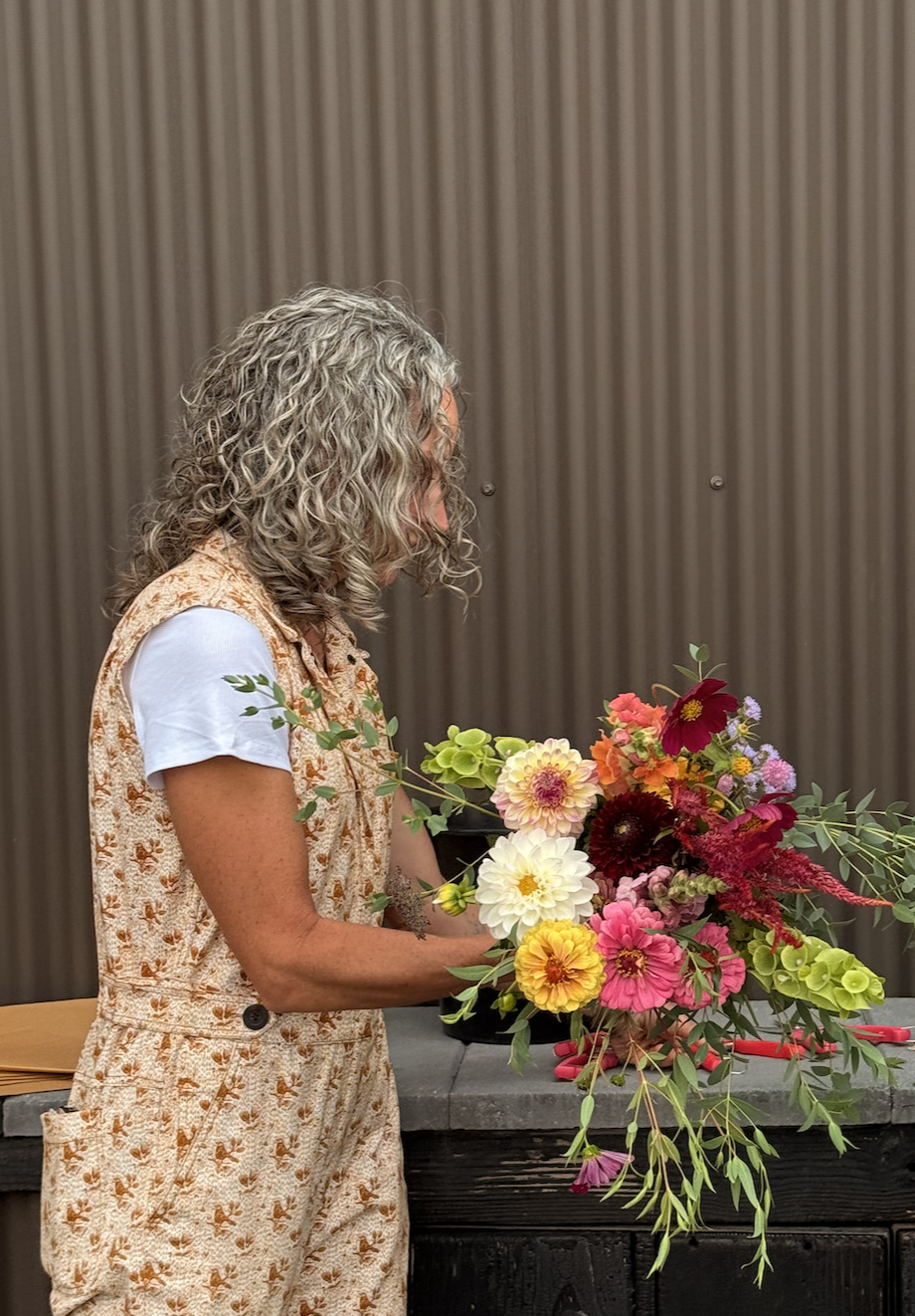 A woman flower farmer with curly grey hair wearing a jumpsuit arranging a color bouquet of fresh flowers including dahlias