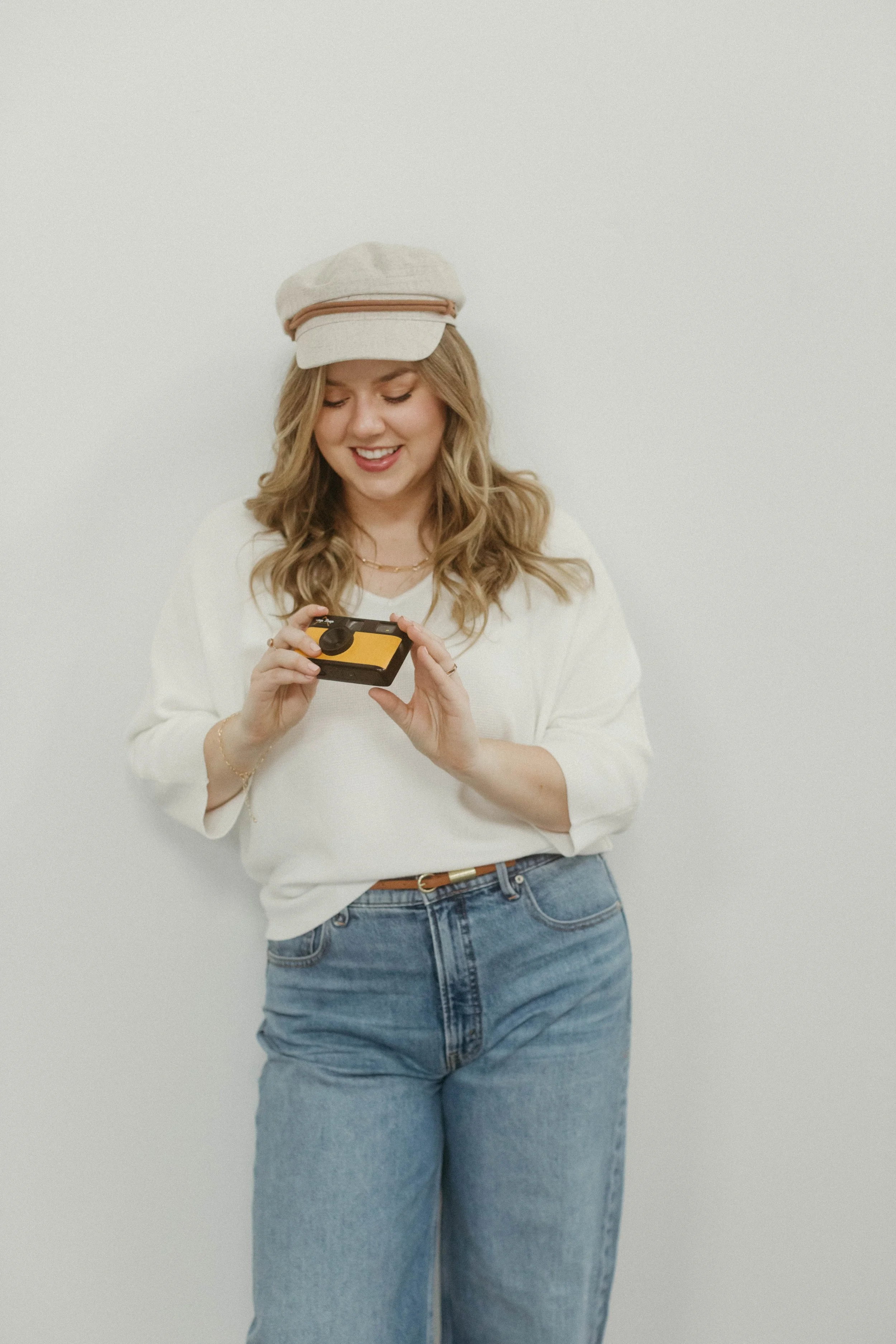 A young woman with wavy blonde hair, wearing a beige cap, white sweater, and blue jeans, looking down and smiling while holding a small electronic device.