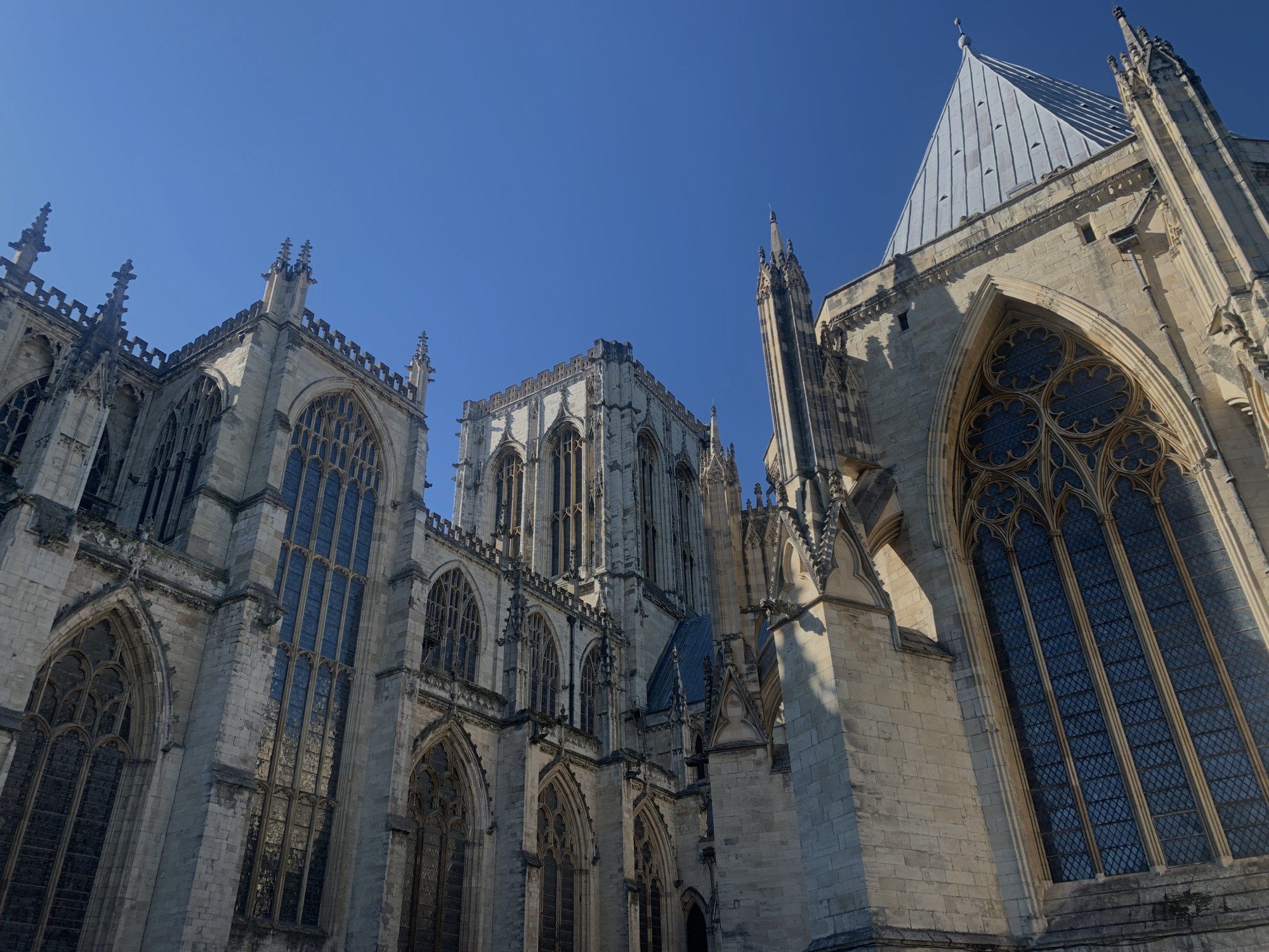 Low-angle view of a Gothic cathedral with large stained glass windows, pointed arches, and intricate stone detailing under a clear blue sky.