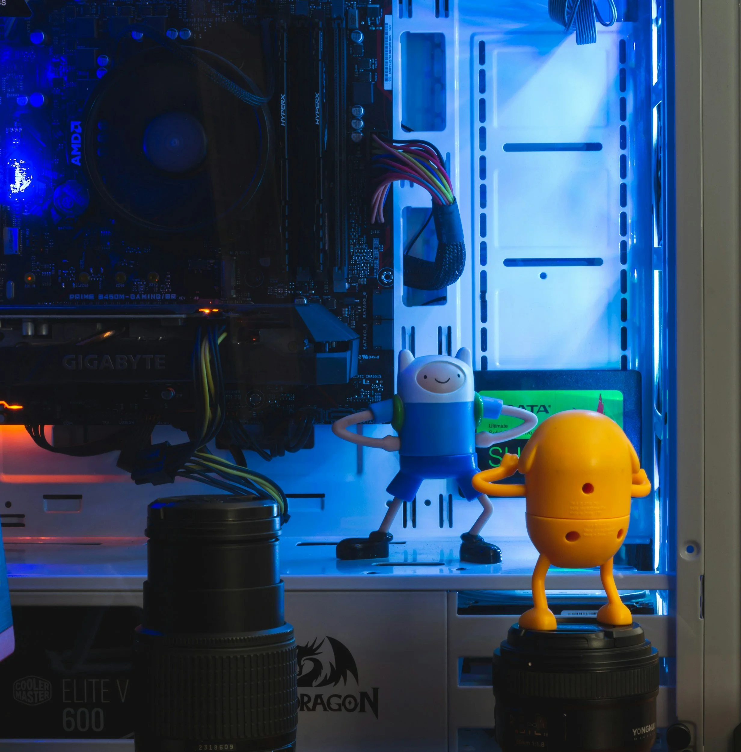 Inside a computer case with blue LED lighting, featuring a motherboard, a CPU cooler, and two cartoon figurines standing on camera lenses, with one resembling a human and the other a yellow, rounded dog-like creature.
