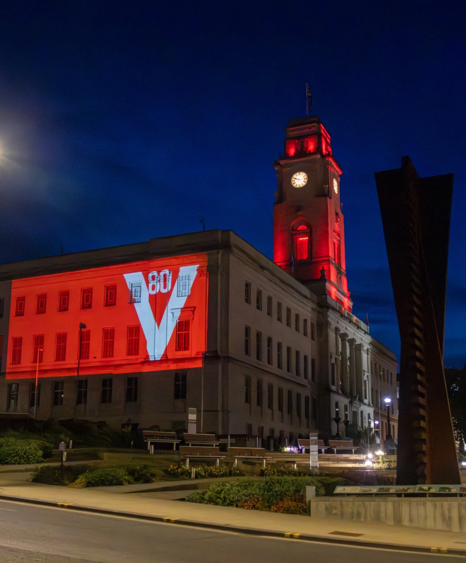 Night-time view of a historic building with a clock tower illuminated in red lights, with a projected logo on the side of the building and sculptures in the foreground.