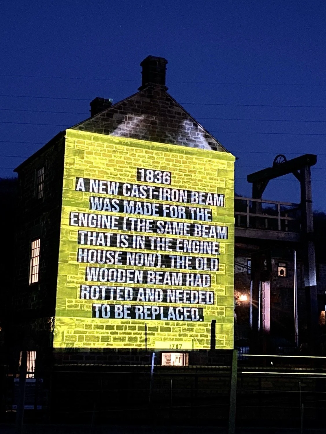A projection of historical text on a stone building at night, describing the replacement of a wooden beam in 1836 with a cast-iron beam for the engine, contrasting old and new materials.