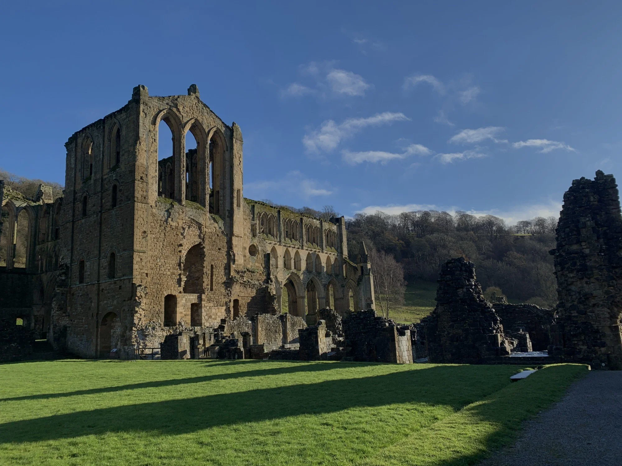 Ruins of an ancient stone building with large arched windows, partially collapsed walls, and a green grassy foreground under a blue sky with some clouds.