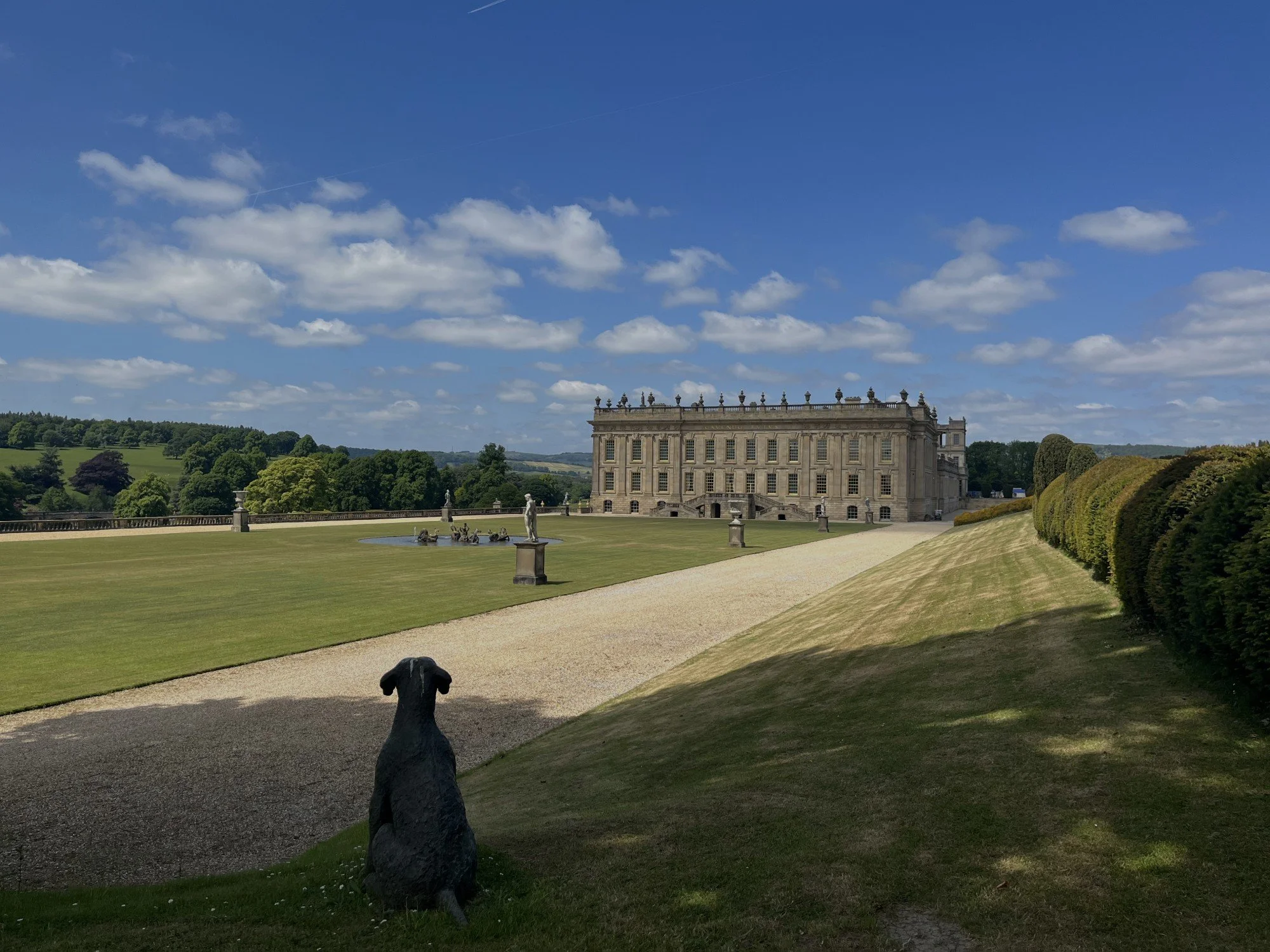 A large historic mansion or palace surrounded by garden grounds, with a statue of a dog in the foreground, and a gravel path leading towards the building, under a partly cloudy blue sky.