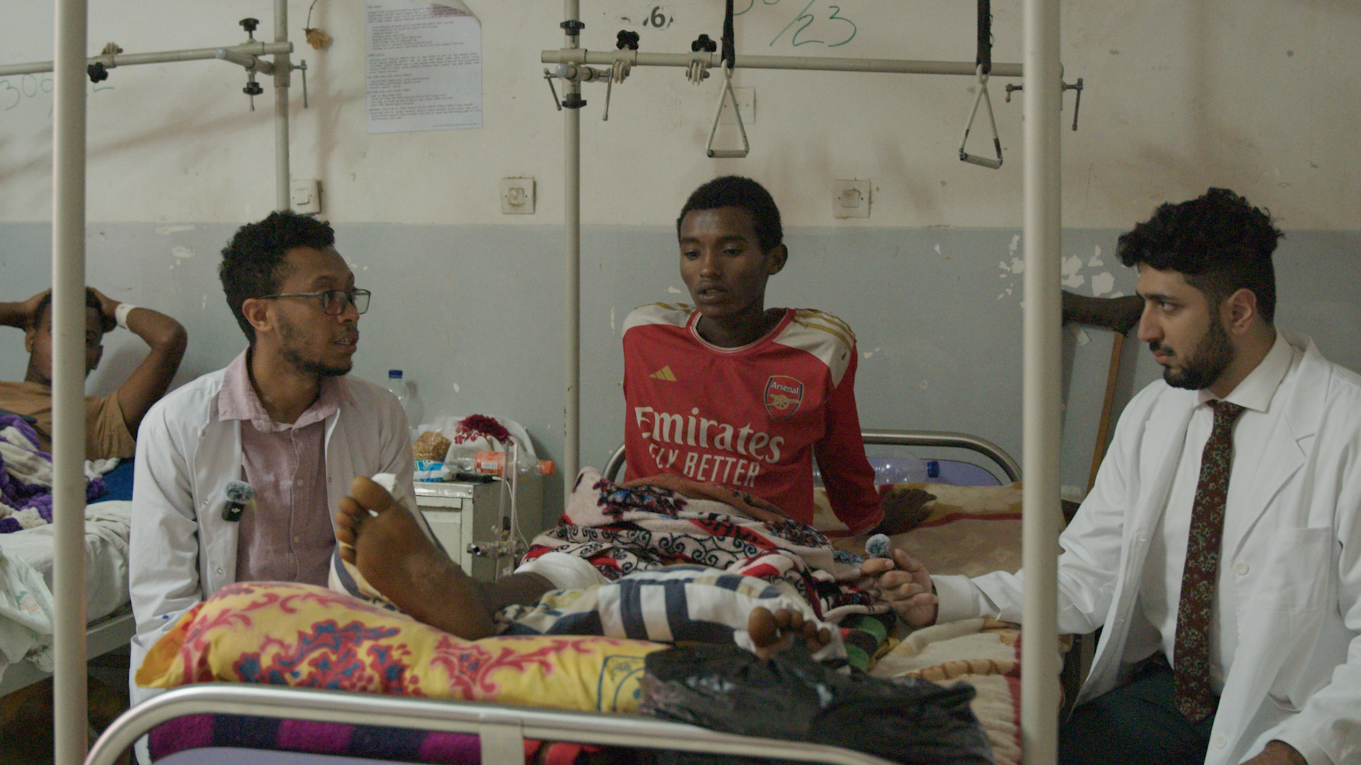A young man in a red Arsenal soccer jersey sits in a hospital bed covered with a blanket, with two men in white coats on either side of him. One man appears to be a doctor, and the other possibly a nurse or medical professional. There are medical equipment and personal items around the room, and another person lies in a bed in the background.