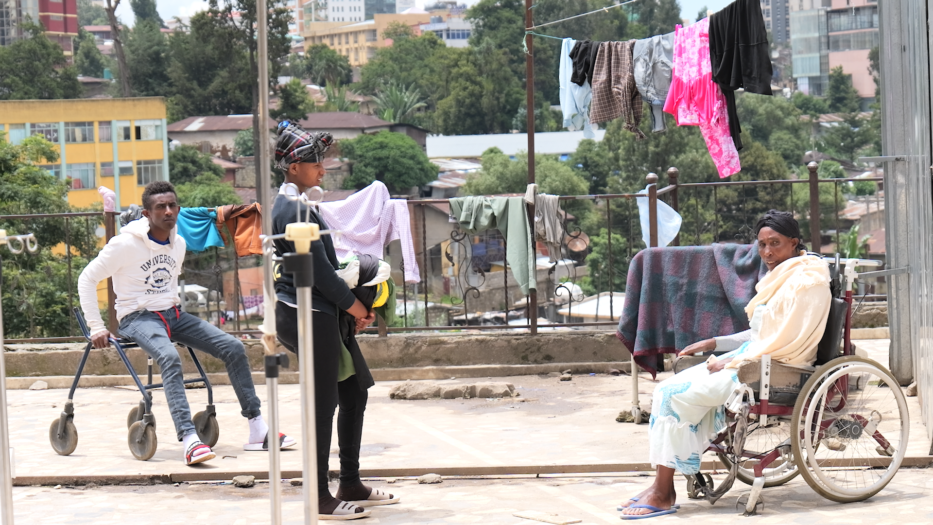 Three individuals, two women and one young man, on a rooftop or outdoor area with laundry hanging on a clothesline, in an urban setting with trees and buildings in the background. One woman is seated in a wheelchair, and the young man is holding a walker.