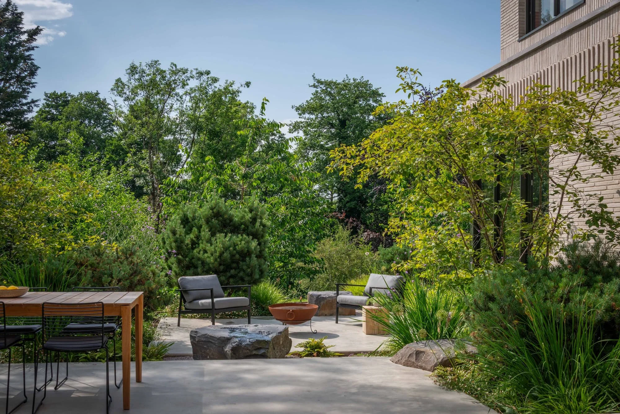 Outdoor patio with concrete floor, wooden dining table, seating area with fire pit, large rocks and water, surrounded by lush trees and plants, contemporary architecture around a modern garden in Ludlow, Shropshire, designed by George William Flynn