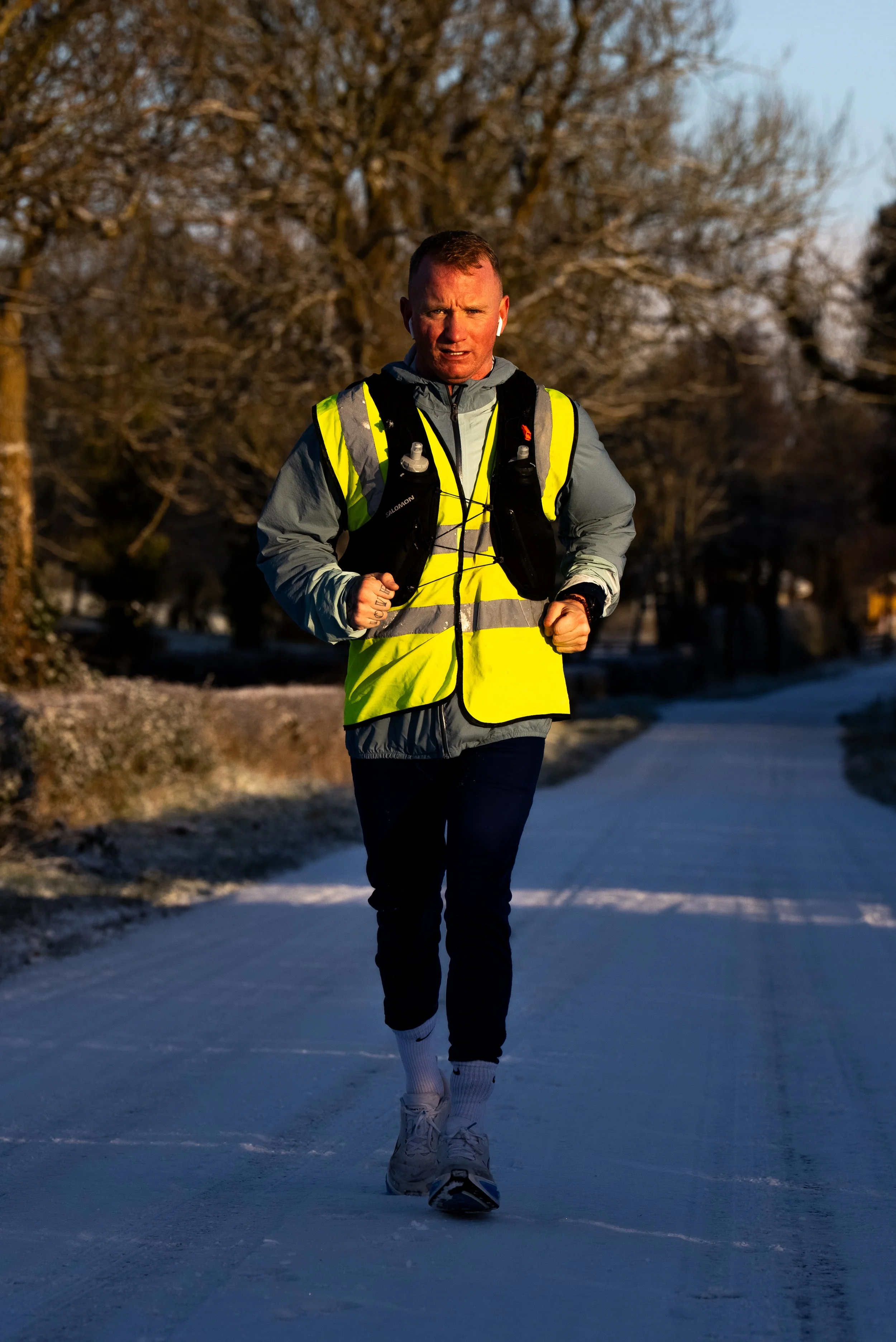 Man running outdoors on a snow-covered path, wearing a reflective yellow vest, gray jacket, black pants, white socks, and running shoes, with trees in the background during sunset.