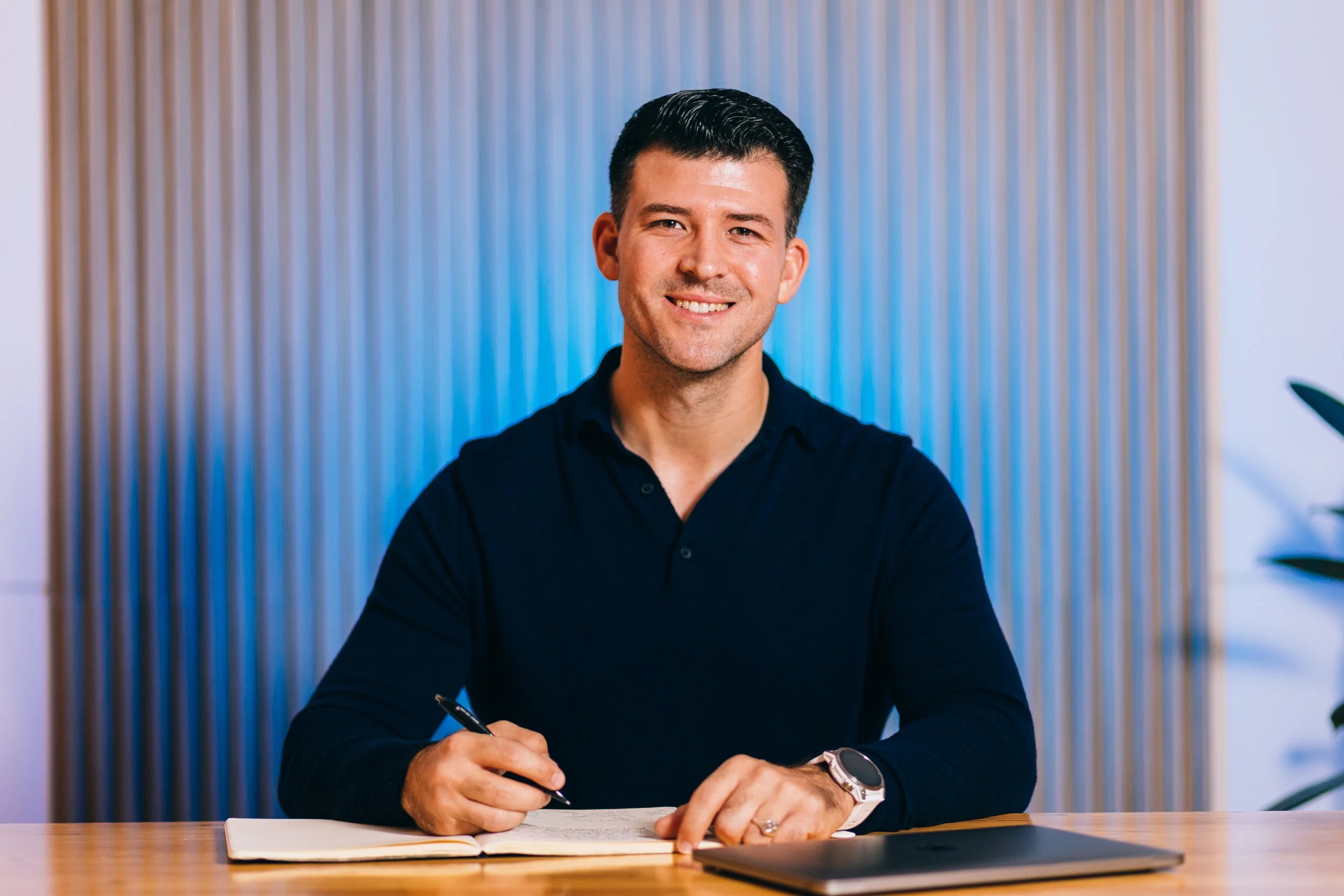 A man sitting at a desk, smiling, holding a pen, with an open notebook, in a modern office setting.