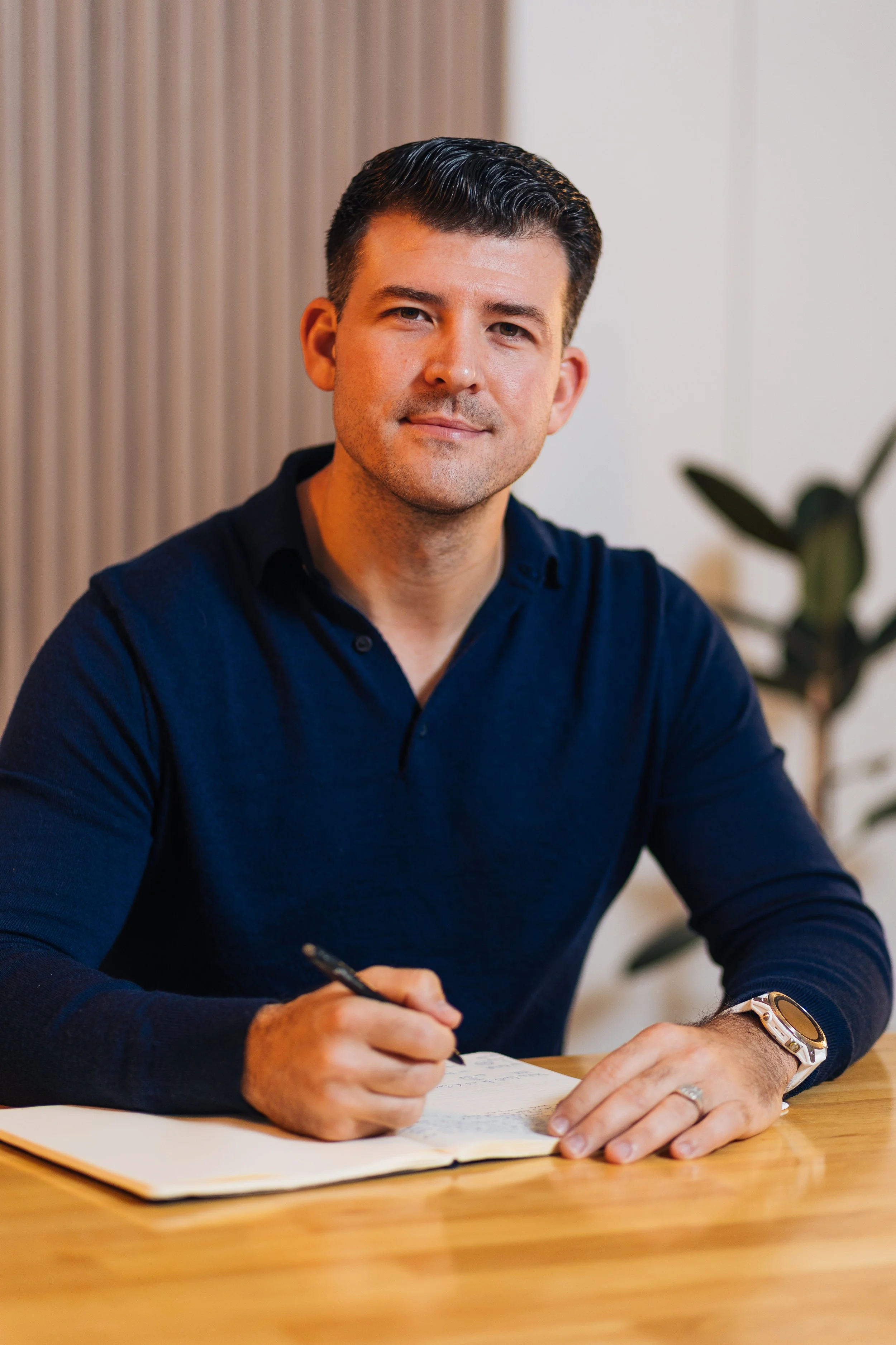 A man with dark hair and a light beard, wearing a dark blue shirt, sitting at a wooden desk, holding a pen, with an open notebook in front of him. There is a plant in the background.