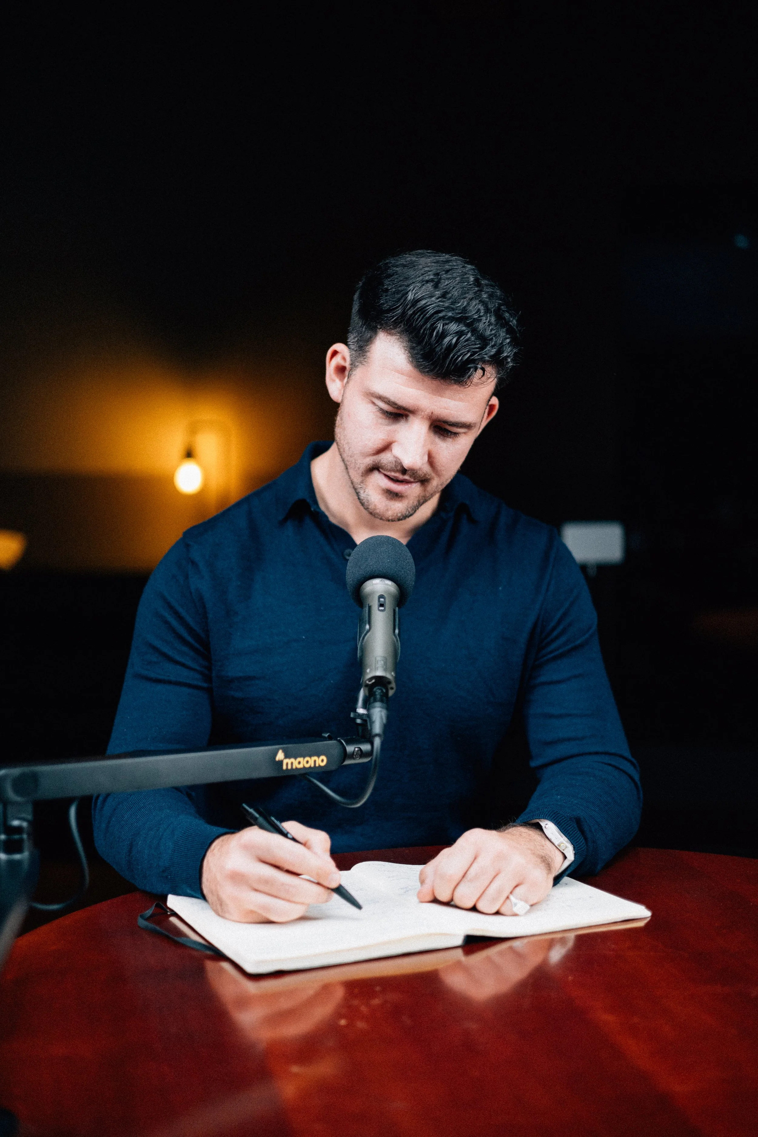 A man with black hair and a beard, wearing a dark blue shirt, sitting at a wooden table with a notebook and pen, recording a podcast with a microphone in front of him, in a dimly lit room.