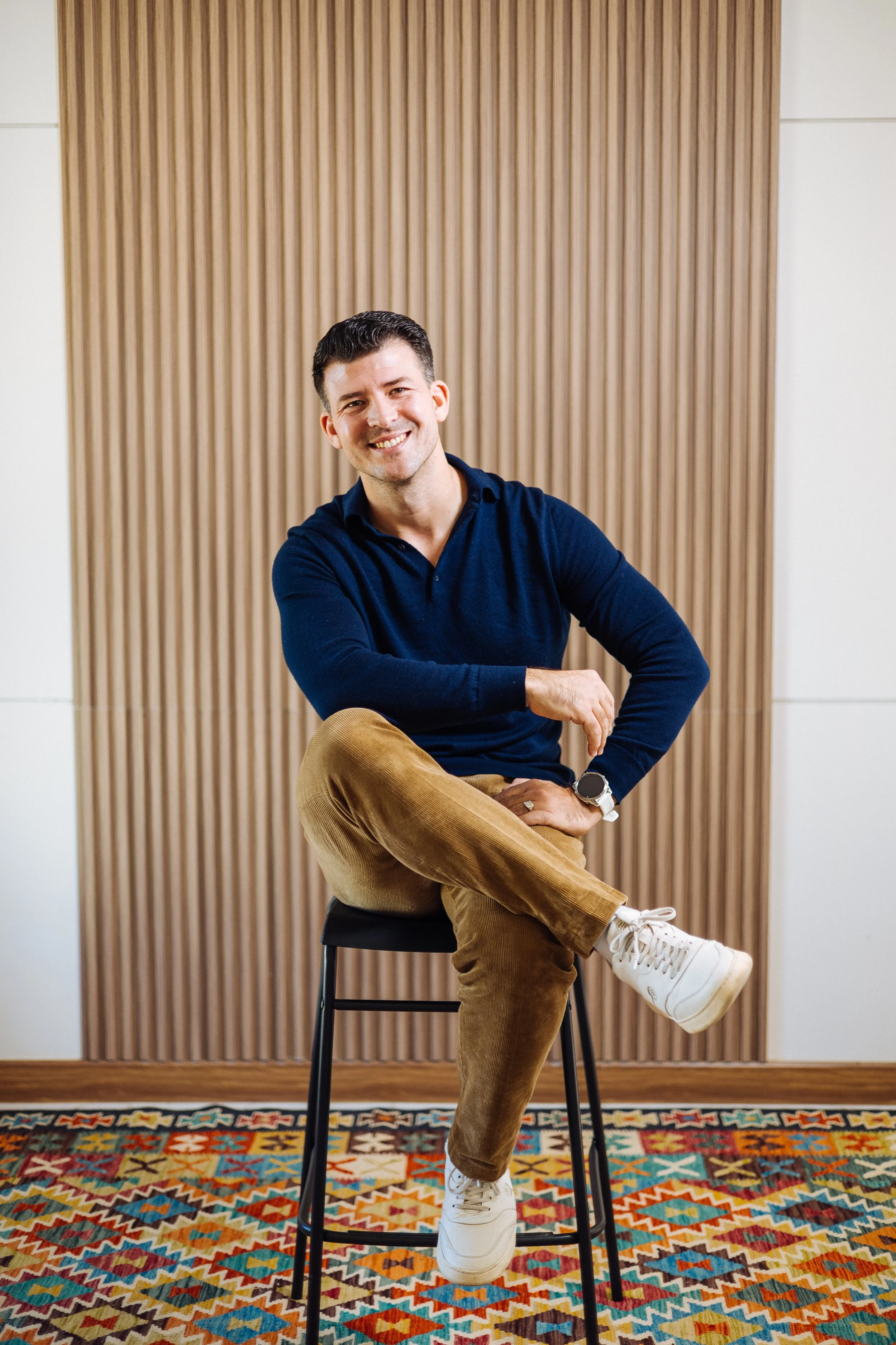 A smiling man sitting on a black stool, wearing a navy blue shirt, tan corduroy pants, white sneakers, and a watch, in front of a wooden wall panel.