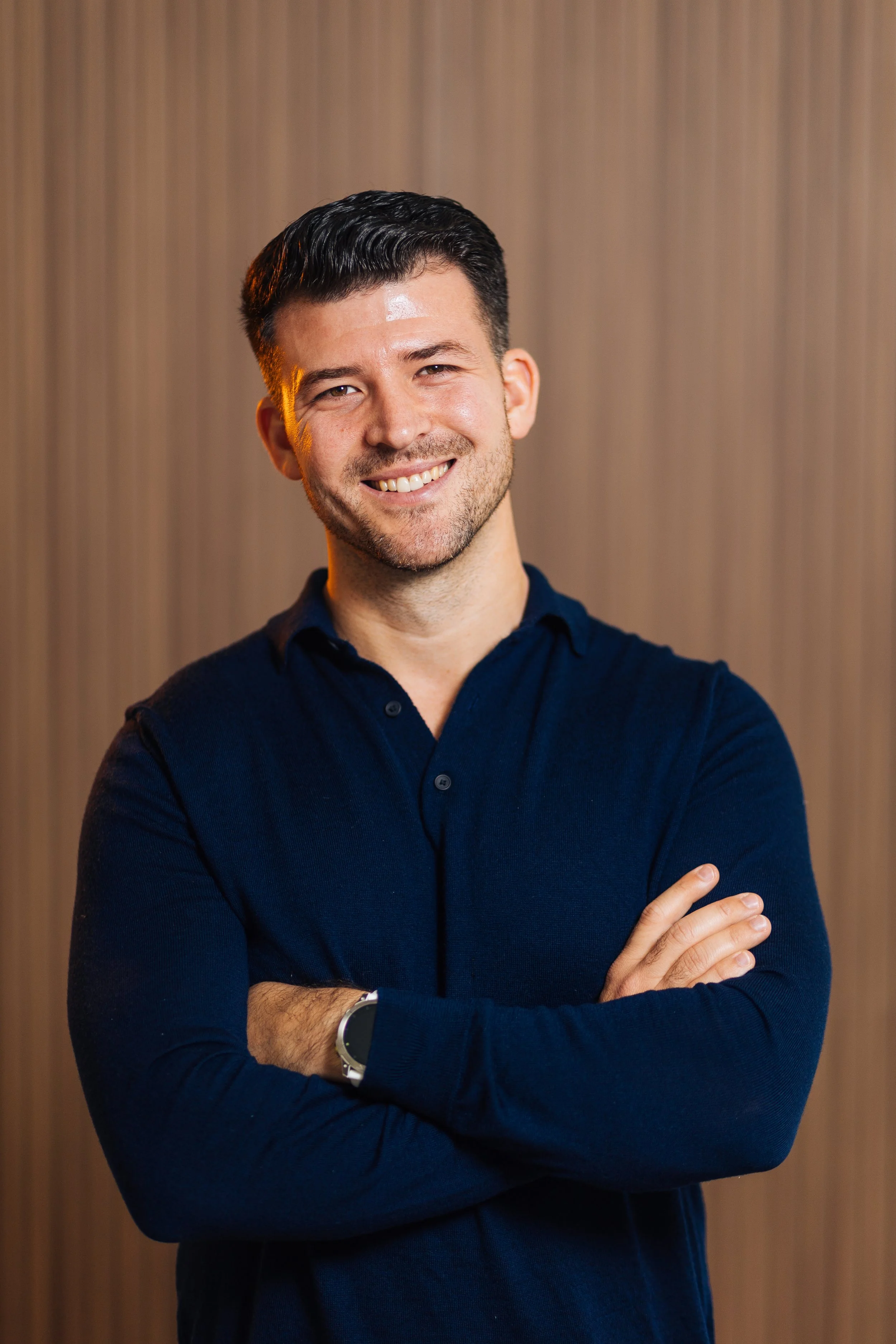 A smiling man with crossed arms, wearing a navy blue shirt and watch, standing against a wood-paneled background.