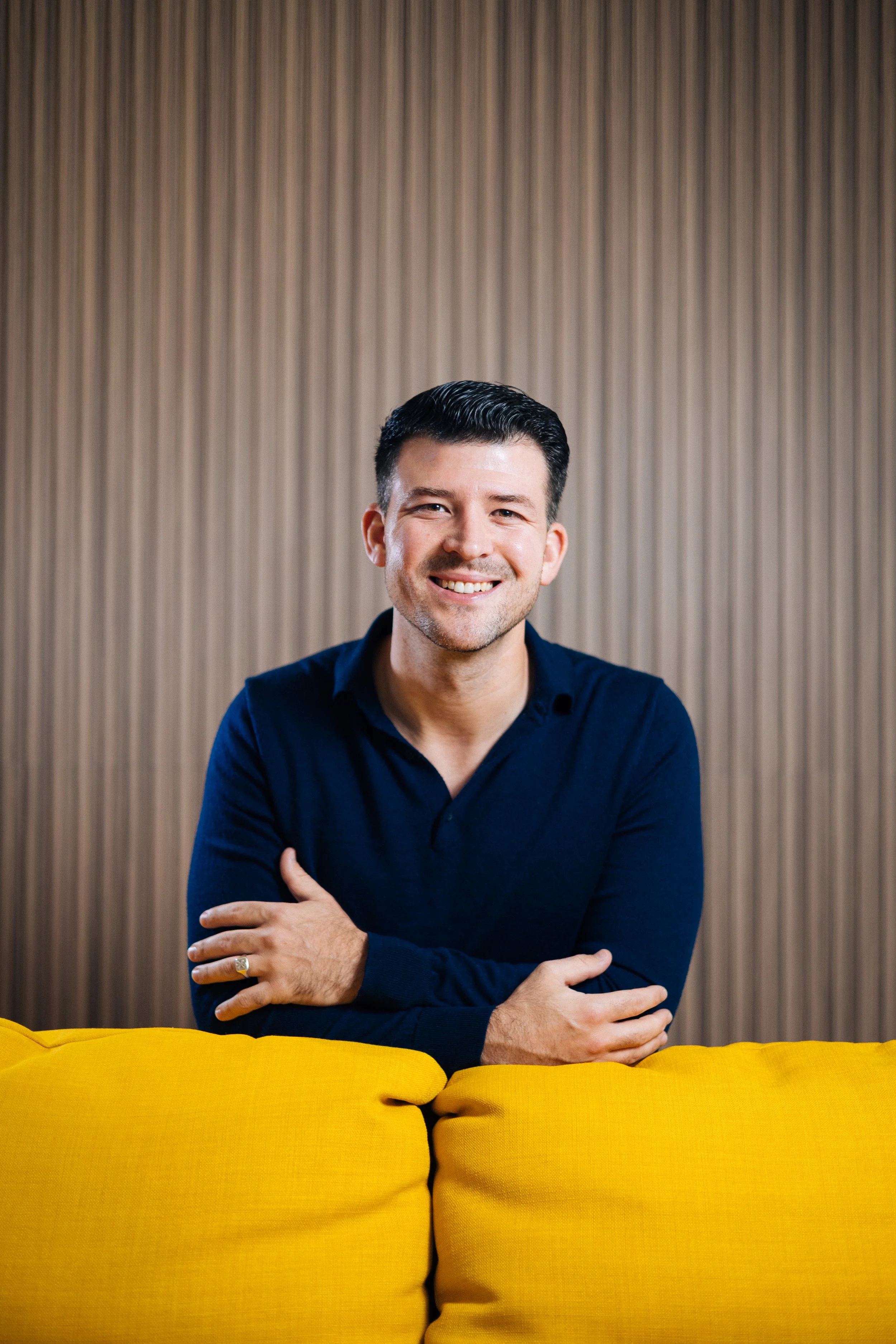 A smiling man with dark hair and a beard, wearing a navy blue shirt, sitting behind a yellow couch with a wooden-paneled wall in the background.