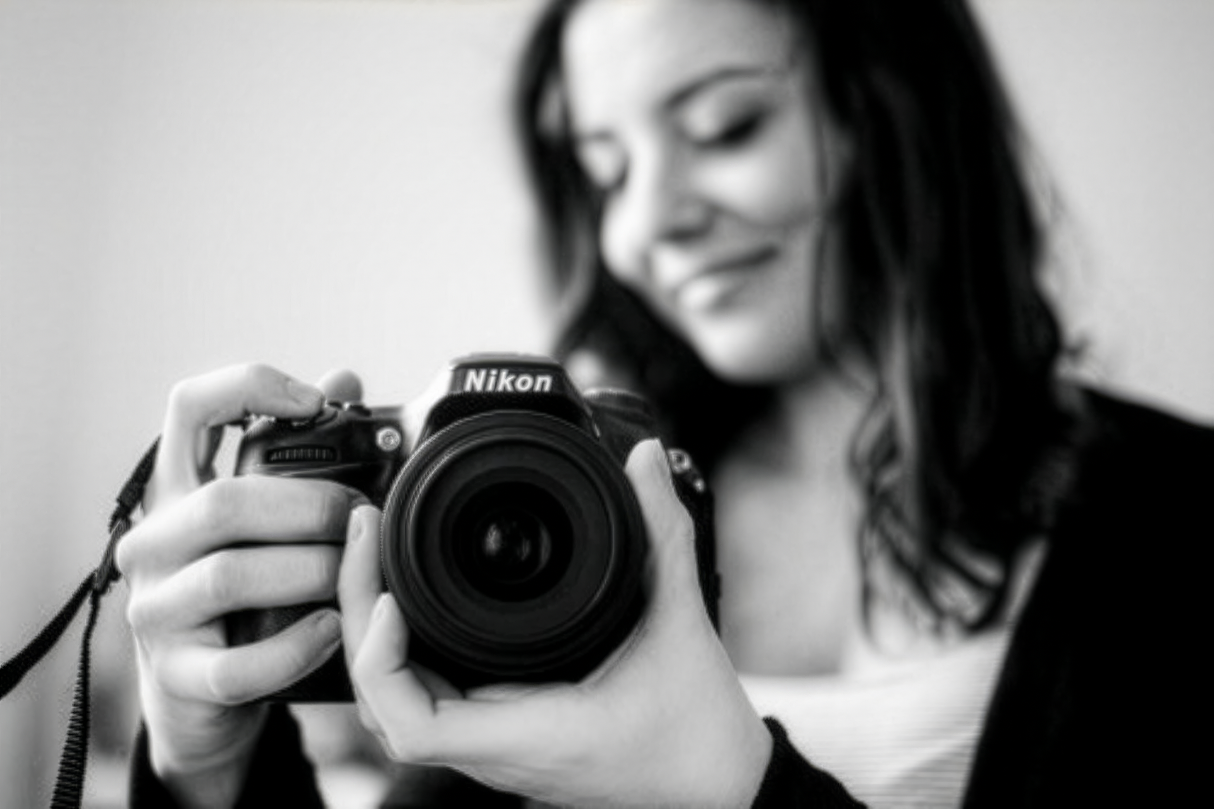 Une femme tient un appareil photo Nikon, en train de se prendre en selfie, sourire, cheveux noirs, habits sombres, fond neutre.