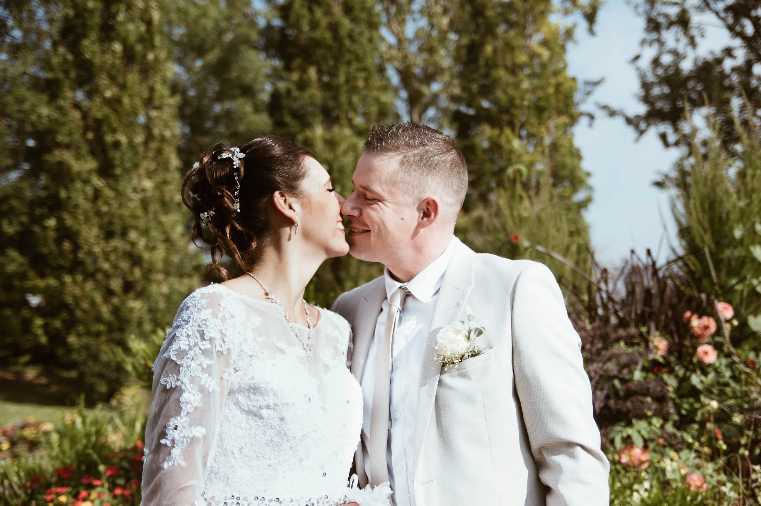 Un couple de mariés en mariage, souriant et se regardant dans un jardin en plein air, avec des arbres et des fleurs en arrière-plan.