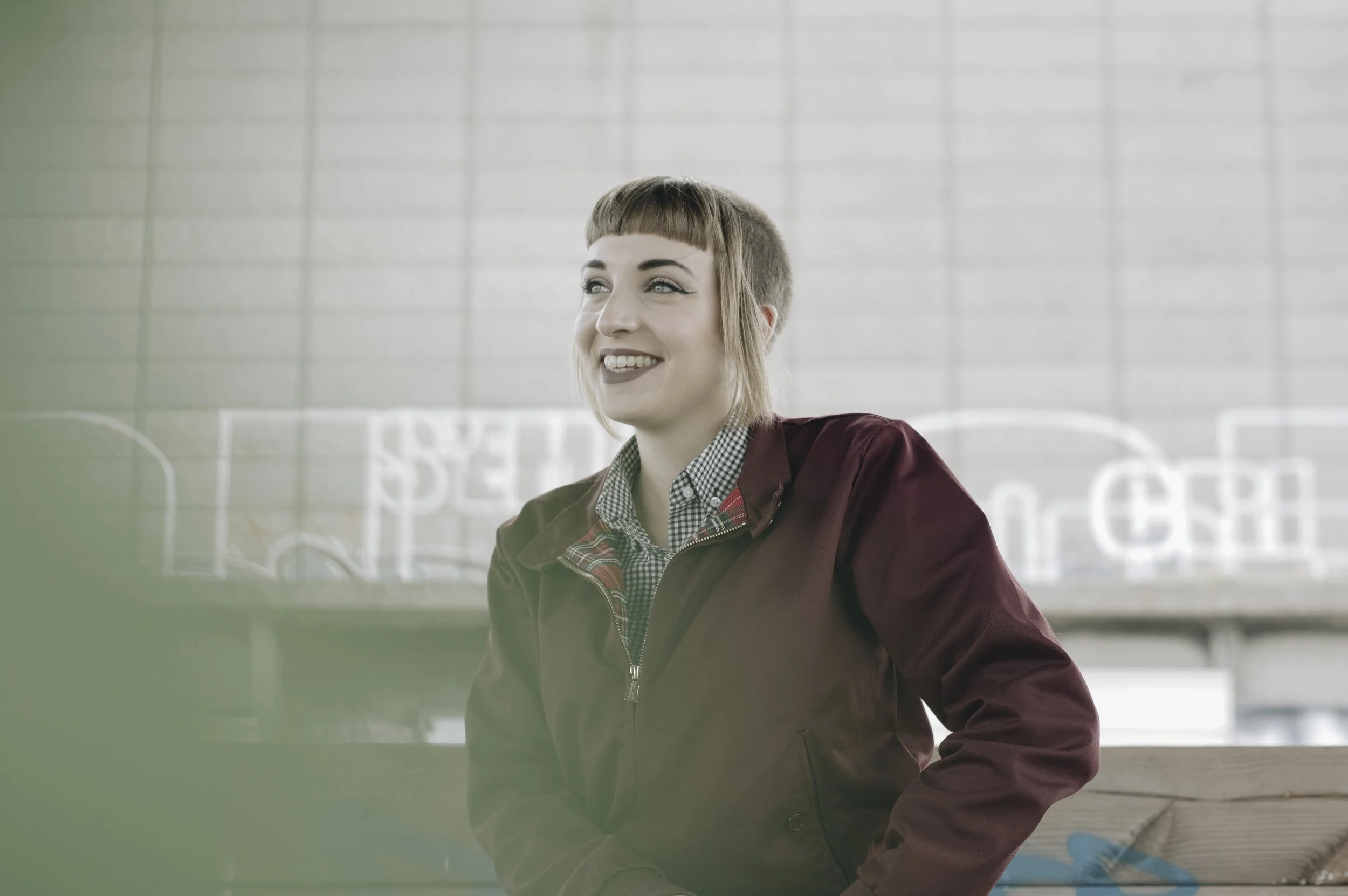 Une jeune femme souriante, avec une coiffure courte et une coupe de cheveux avec une frange, porte une veste marron et un chemisier à carreaux, assise contre un mur en briques avec des graffitis en arrière-plan.