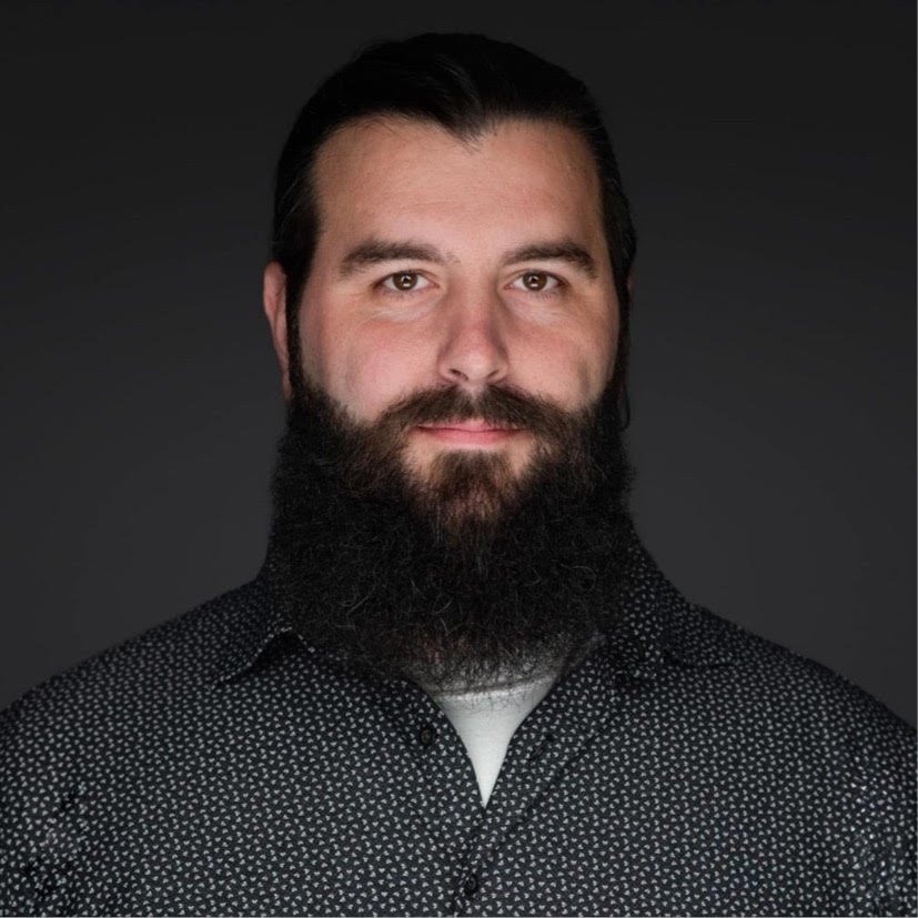 Portrait of a man with dark hair and beard, wearing a black button-up shirt with a white undershirt against a dark background.