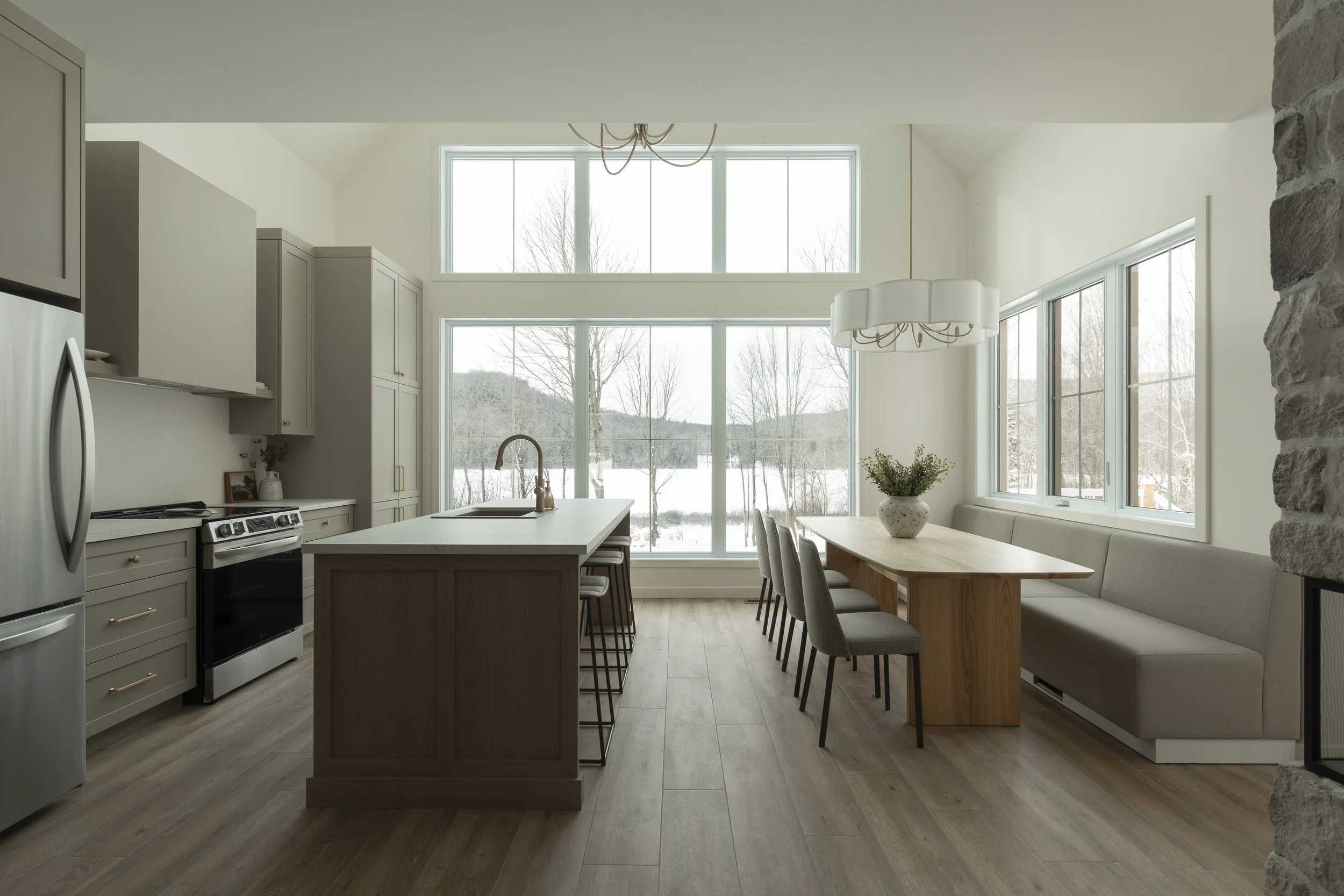 Cuisine lumineuse avec grandes fenêtres donnant sur un paysage enneigé, îlot central avec évier, table en bois avec chaises et banquette intégrée, décor moderne et minimaliste.