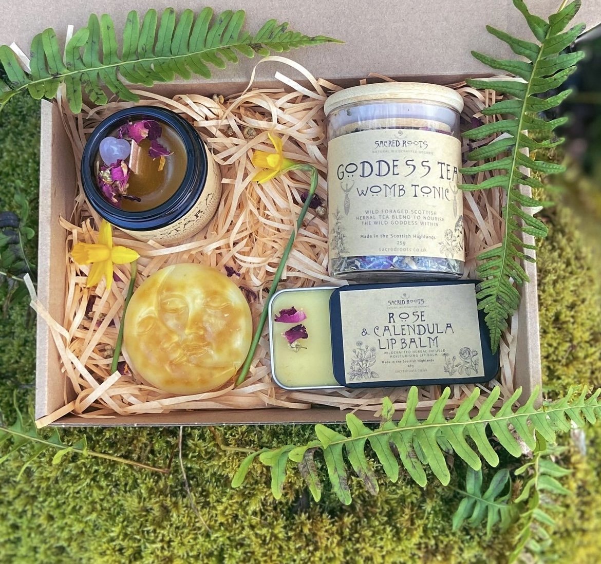 A gift box with herbal wellness products, including a candle with dried flowers, a jar of Goddess Tea Womb Tonic, a tin of Rose & Calendula Lip Balm, and decorative yellow flowers, all arranged on shredded paper and framed by green fern leaves.