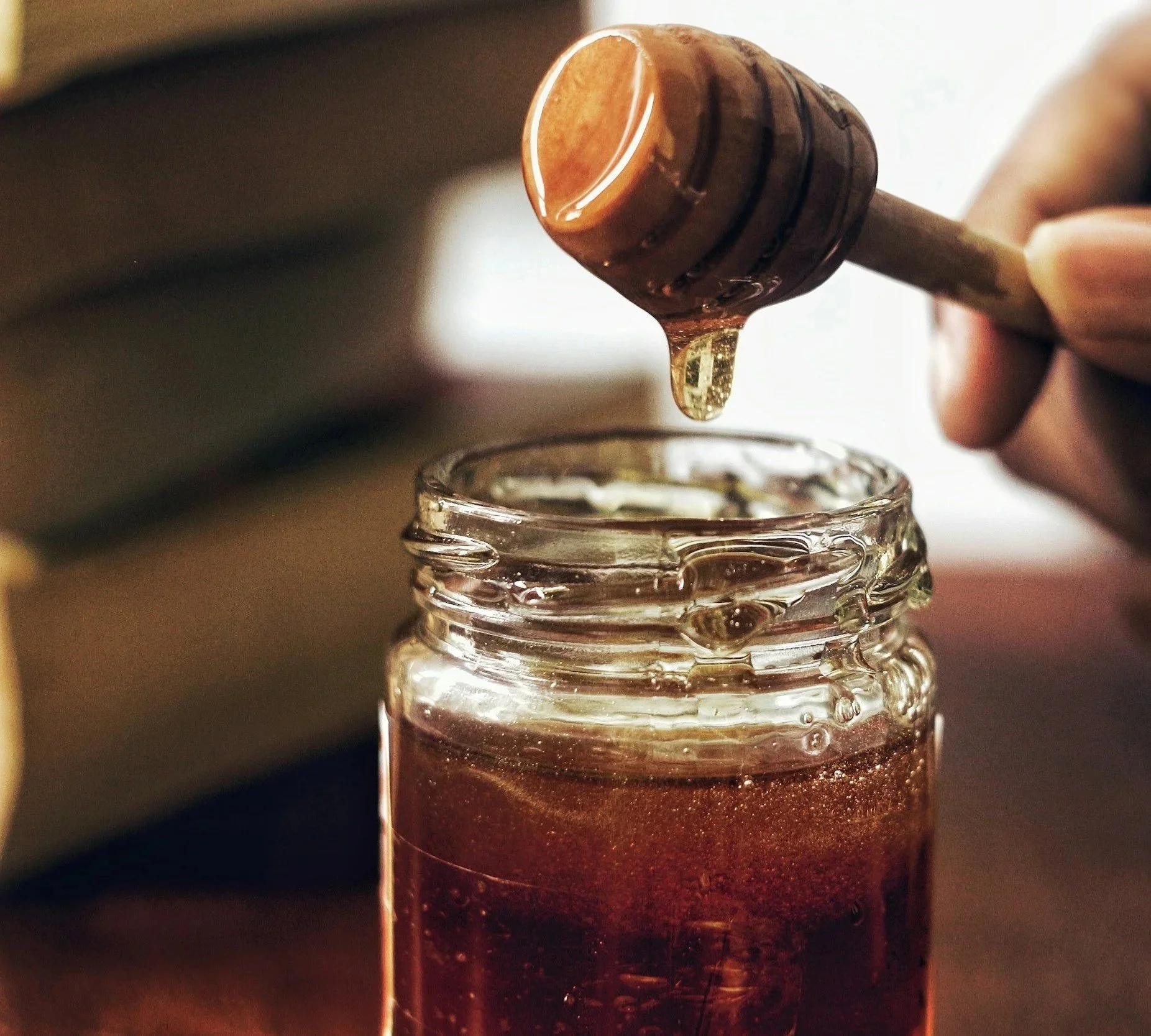 Close-up of honey being drizzled from a honey dipper into a glass jar filled with honey.