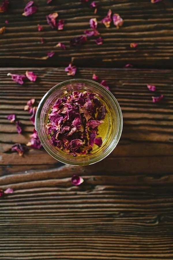 A small glass container with dried pink rose petals on top of a wooden surface, some petals scattered around the container.
