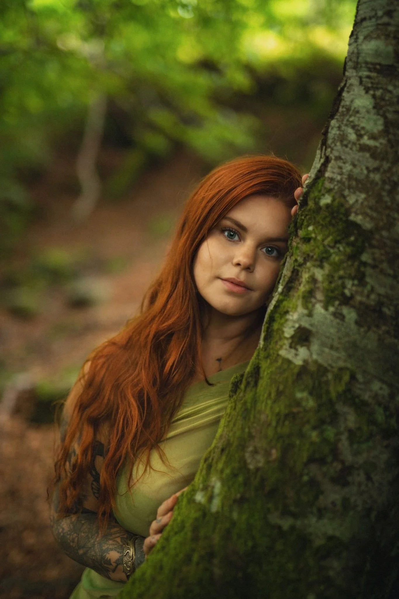 A young woman with long red hair and tattoos on her arm peeking from behind a moss-covered tree in a forest.