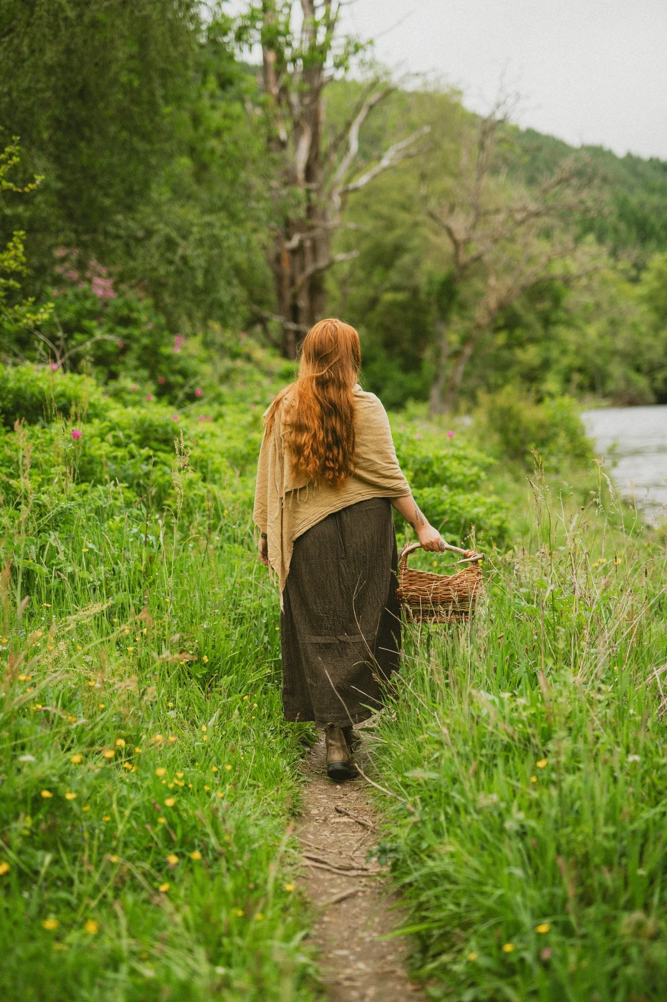 A woman with long red hair, wearing a beige shawl and a long black skirt, walking down a narrow dirt path through a lush green area with tall grass and wildflowers, carrying a wicker basket, by a river.